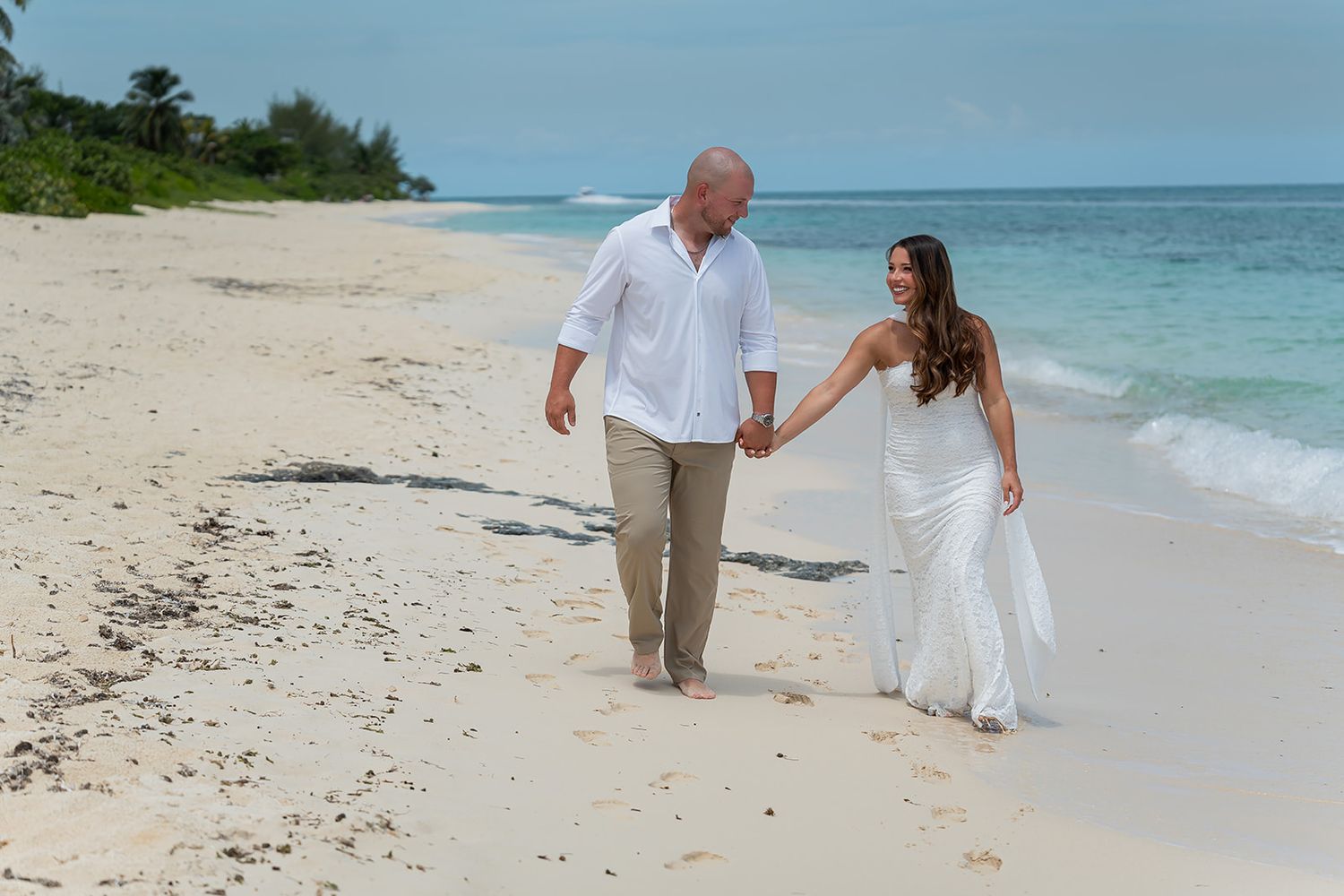 Couple in wedding attire walking hand-in-hand on a tropical white sand beach with turquoise ocean waves.