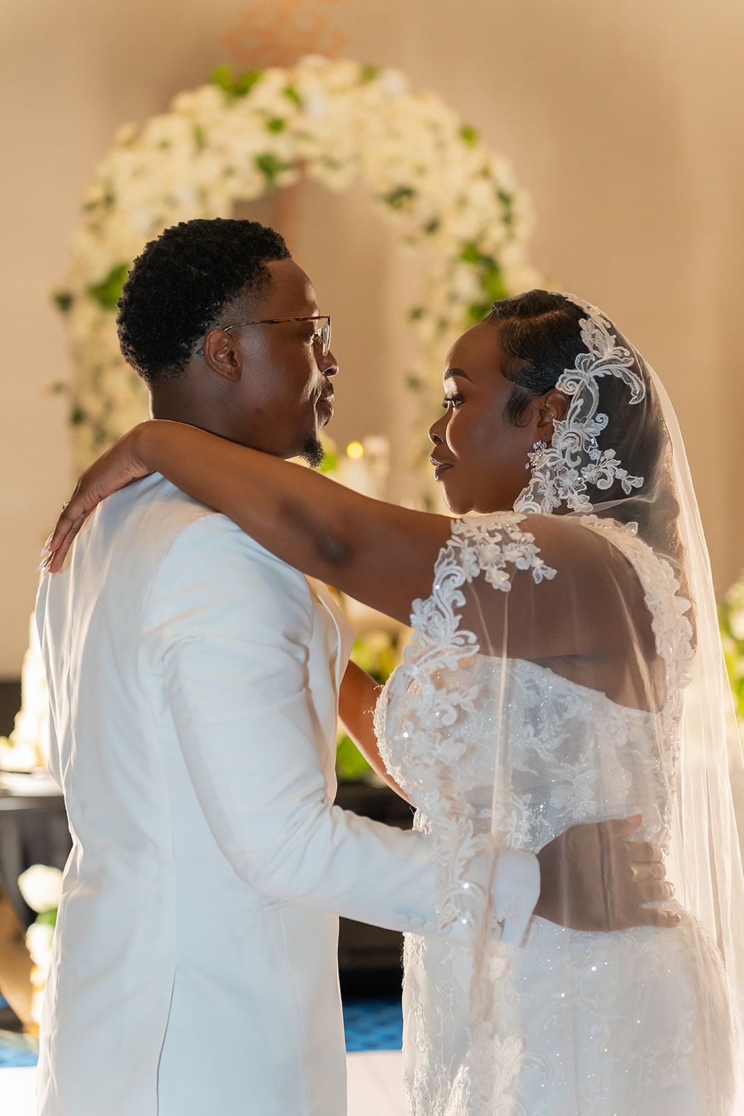 A bride and groom share a tender first dance in elegant white wedding attire before a floral arch.