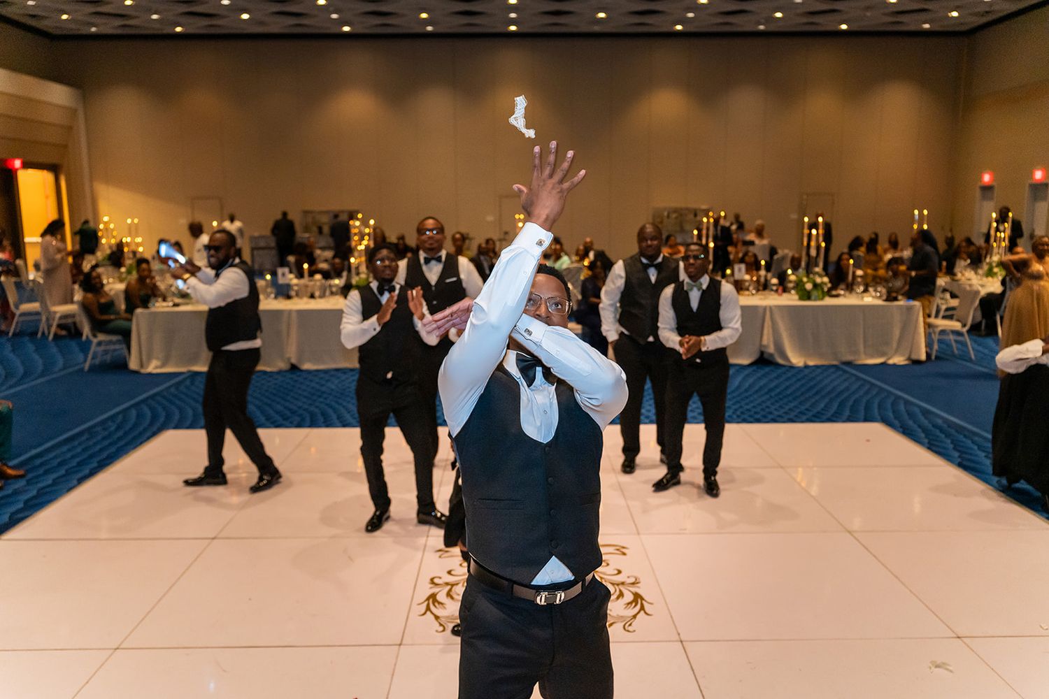 Groom tosses boutonniere to groomsmen on white dance floor at elegant wedding reception venue.