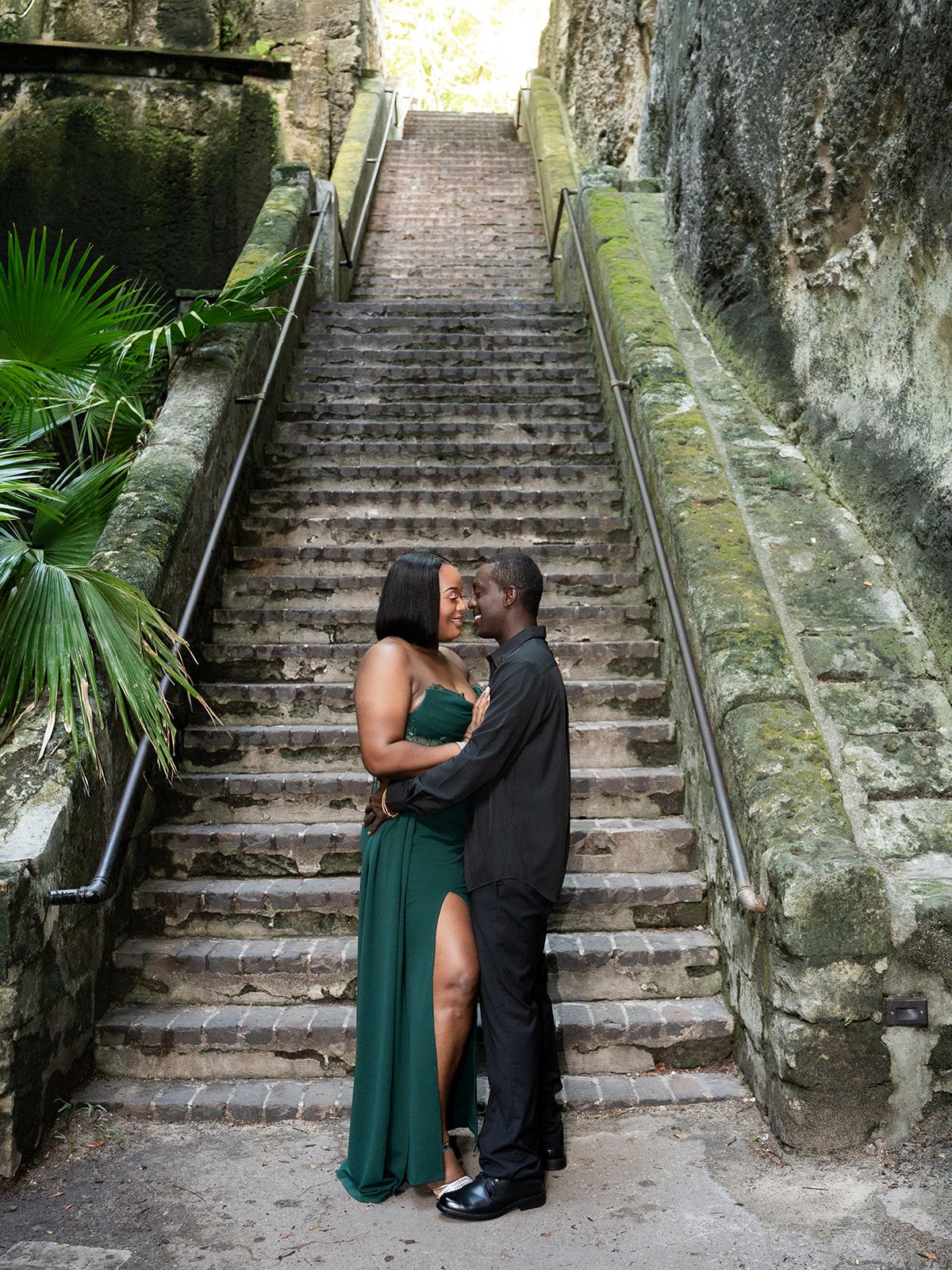 Couple in formal wear embracing and kissing on historic stone staircase surrounded by lush greenery.