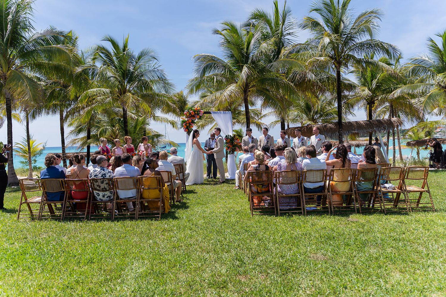 Outdoor beach wedding ceremony with guests seated on chairs surrounded by palm trees and ocean views.