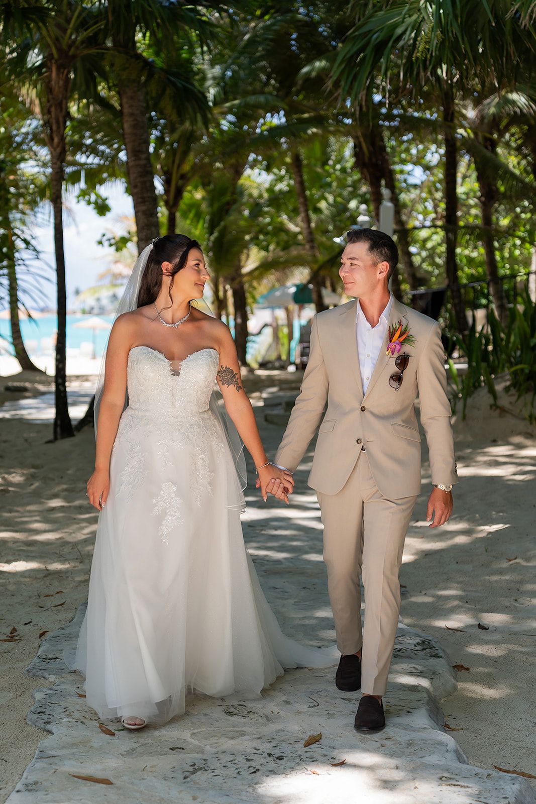 Bride and groom walking hand in hand on a tropical beach path surrounded by palm trees in wedding attire.