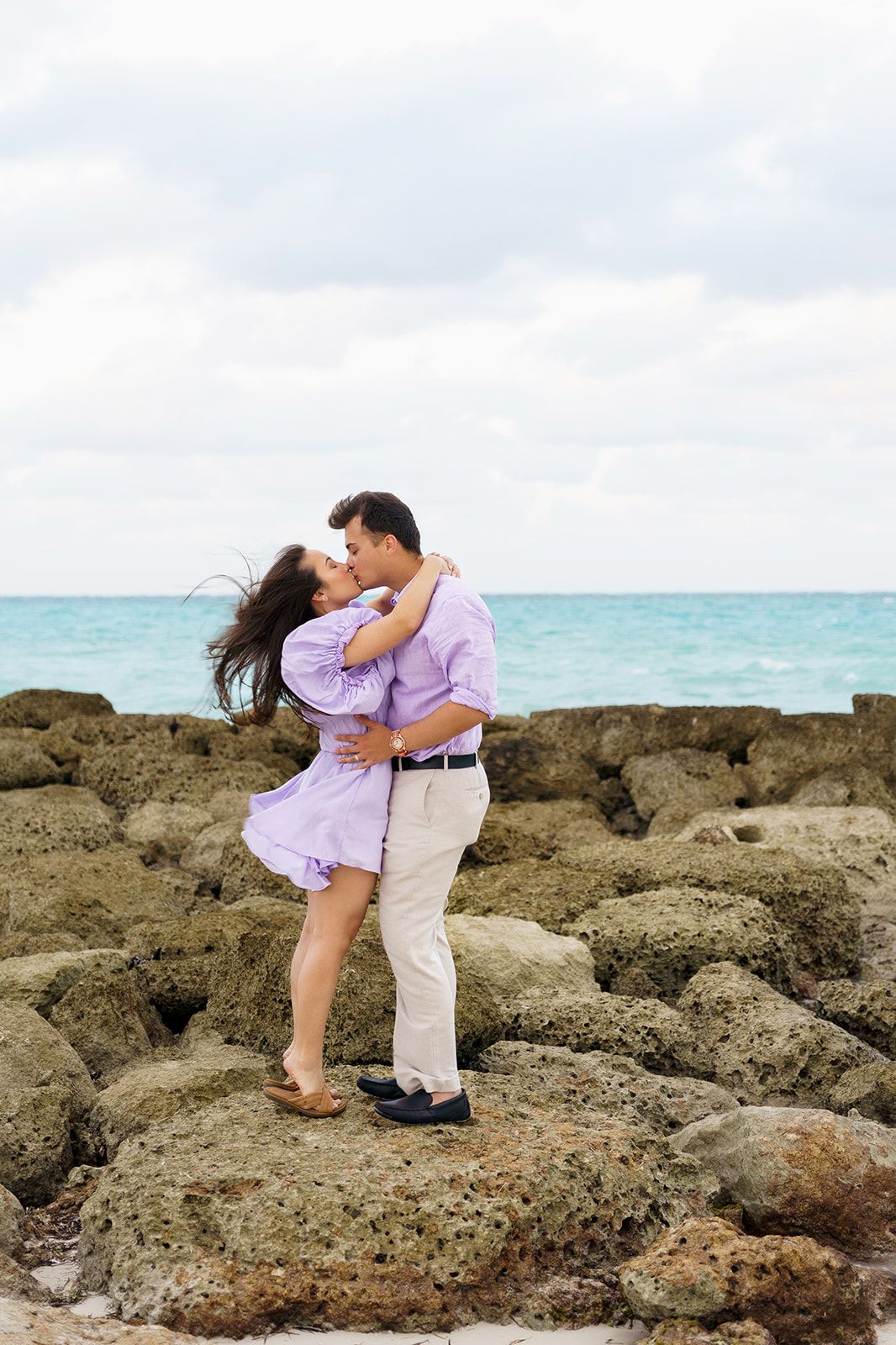 Couple embracing and kissing on rocky beach with turquoise ocean backdrop on a cloudy day.
