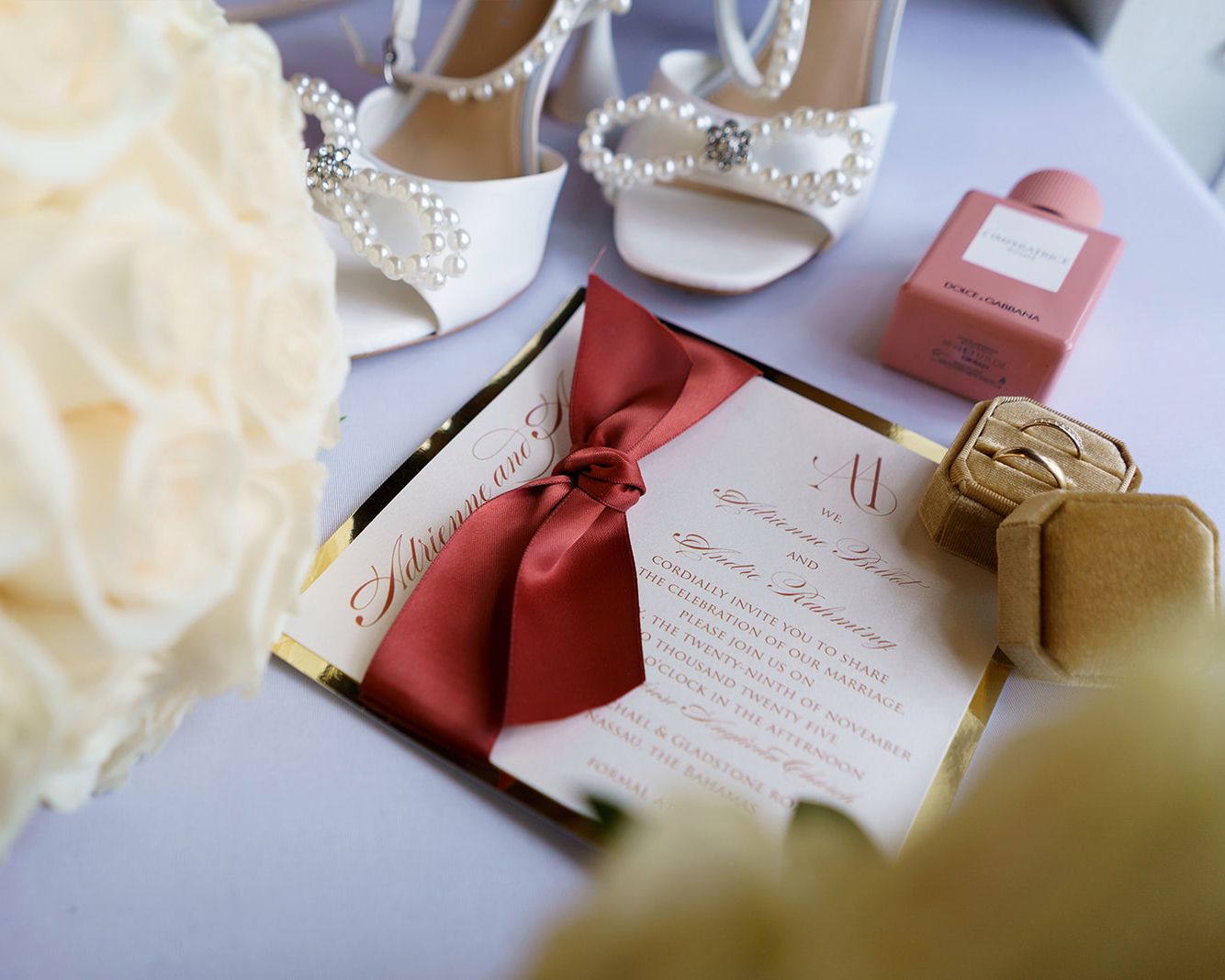 Elegant wedding flat lay with pearl heels, red ribbon invitation, floral bouquet, and luxury perfume bottle.