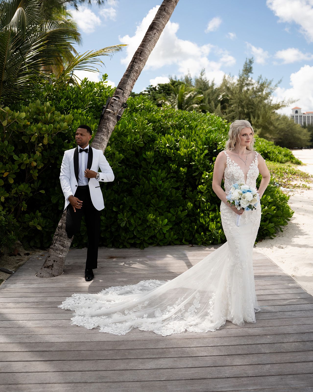 Bride in lace mermaid gown holds bouquet on wooden boardwalk as groom in white tuxedo leans on palm tree.