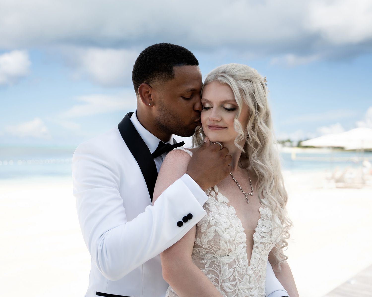 Interracial couple shares tender kiss on tropical beach in elegant wedding attire.