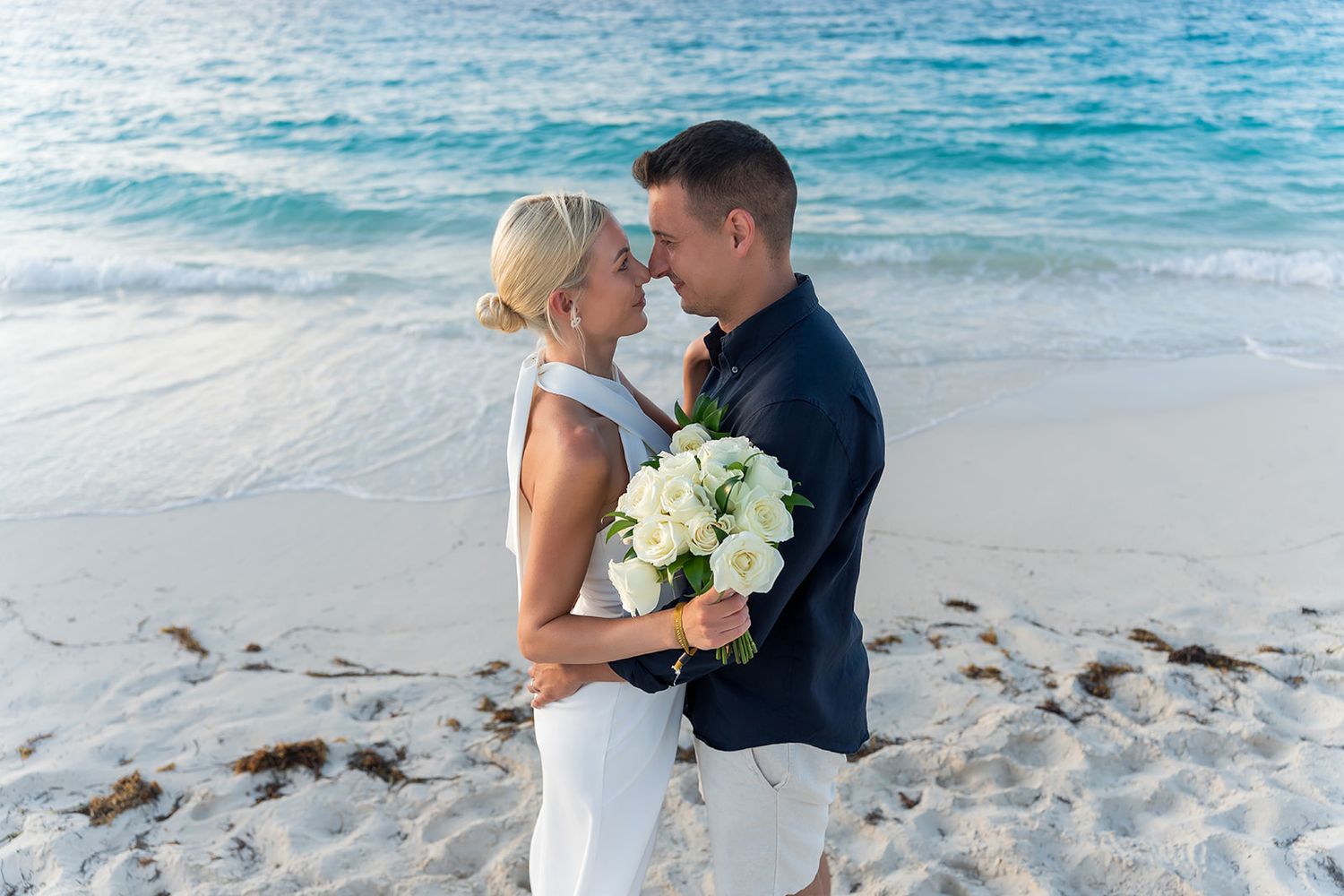 Newlyweds sharing a romantic moment on a white sand beach, bride holding white flower bouquet.