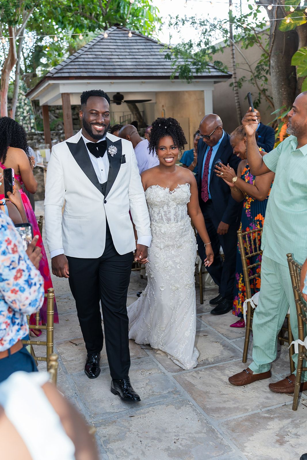 Smiling newlywed couple walks down outdoor wedding aisle, groom in white tuxedo jacket, bride in beaded strapless gown.