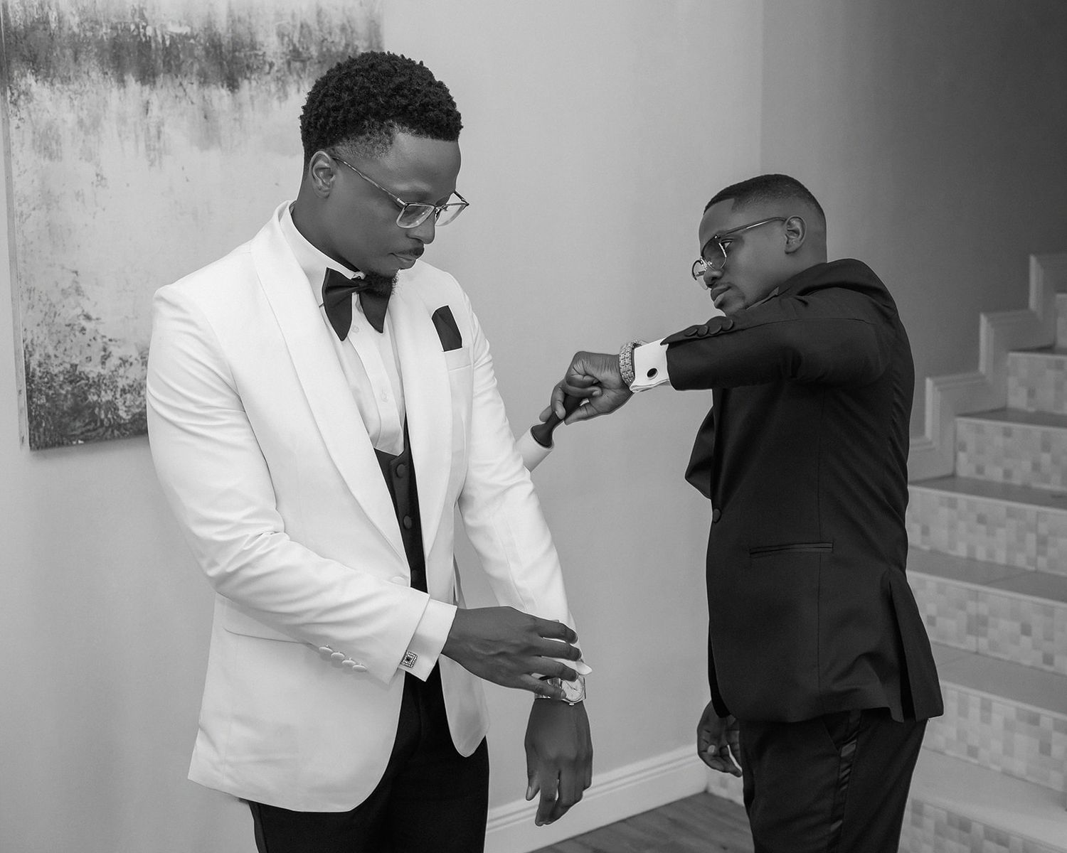 Two men in formal suits fist bumping, one in white tuxedo jacket, in a black and white photo.