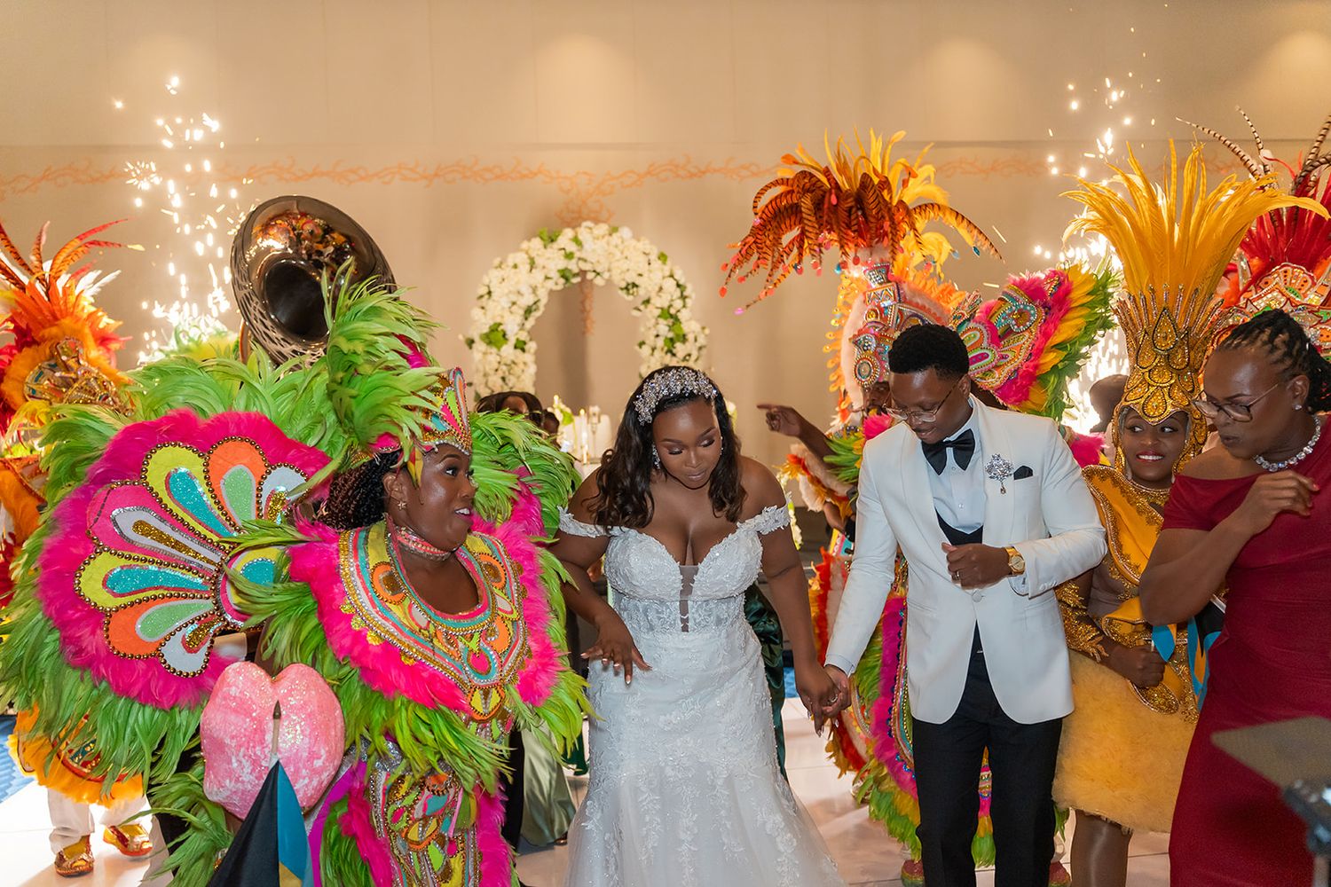 Bride and groom celebrate with colorful Junkanoo performers in feathered costumes at festive wedding.