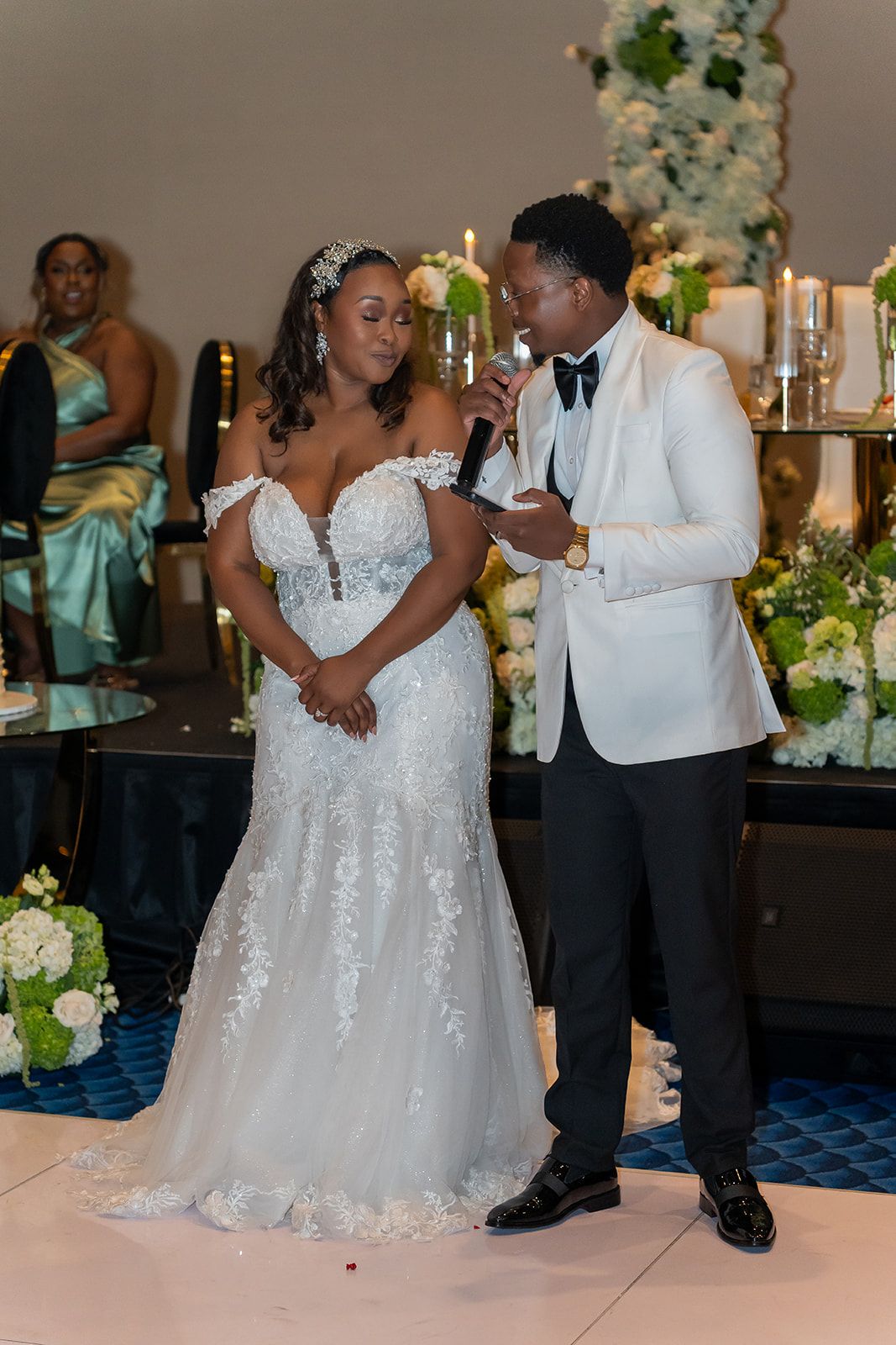 Bride in lace mermaid gown and groom in white tuxedo share a speech at their elegant wedding reception.