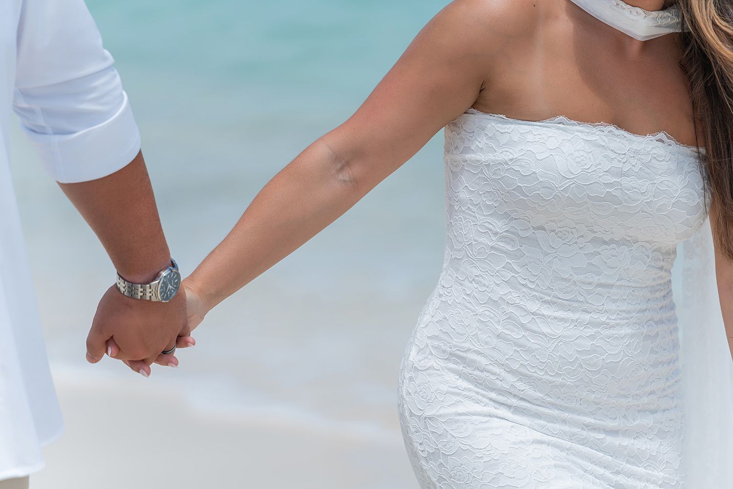 Couple holding hands on beach, bride in white strapless lace dress, groom in white shirt, ocean background.