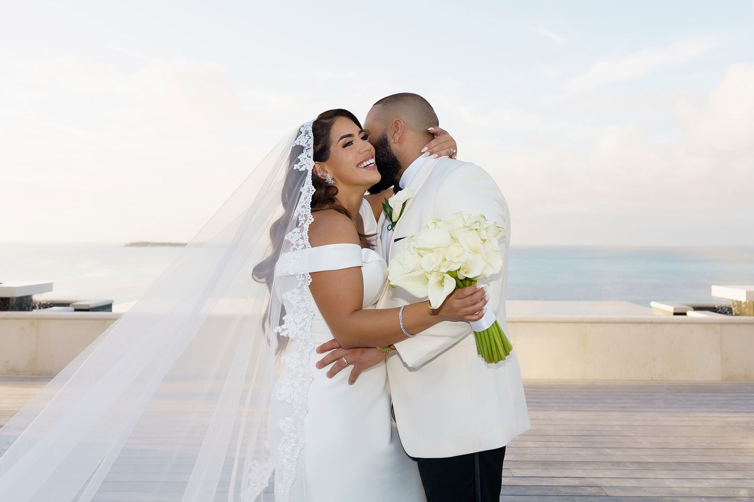 Bride and groom embrace on rooftop terrace, she holds white bouquet wearing veil, he wears white tuxedo jacket, ocean backdrop.