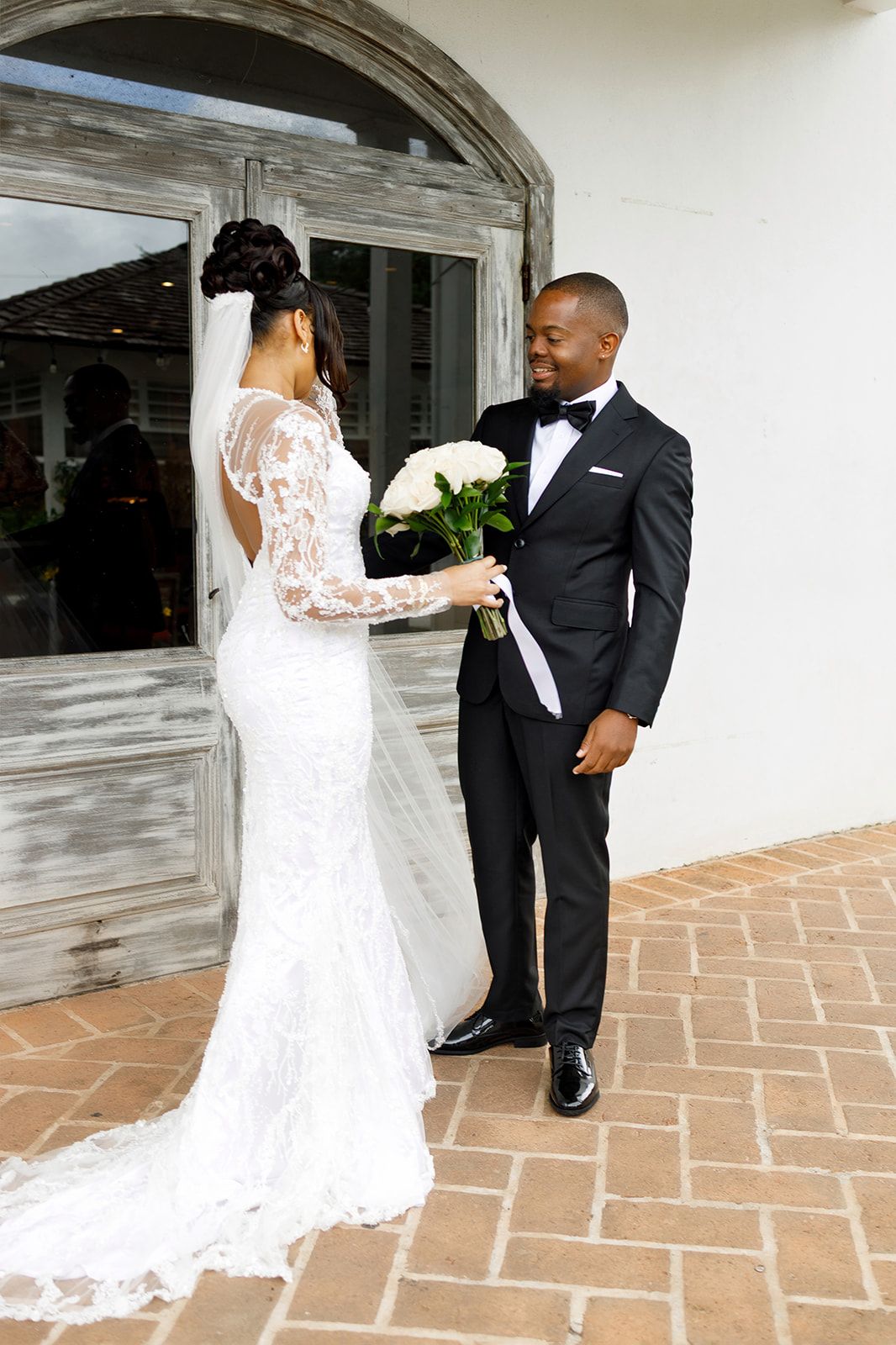 Bride in lace gown and groom in black tuxedo share a romantic moment outside a rustic venue.