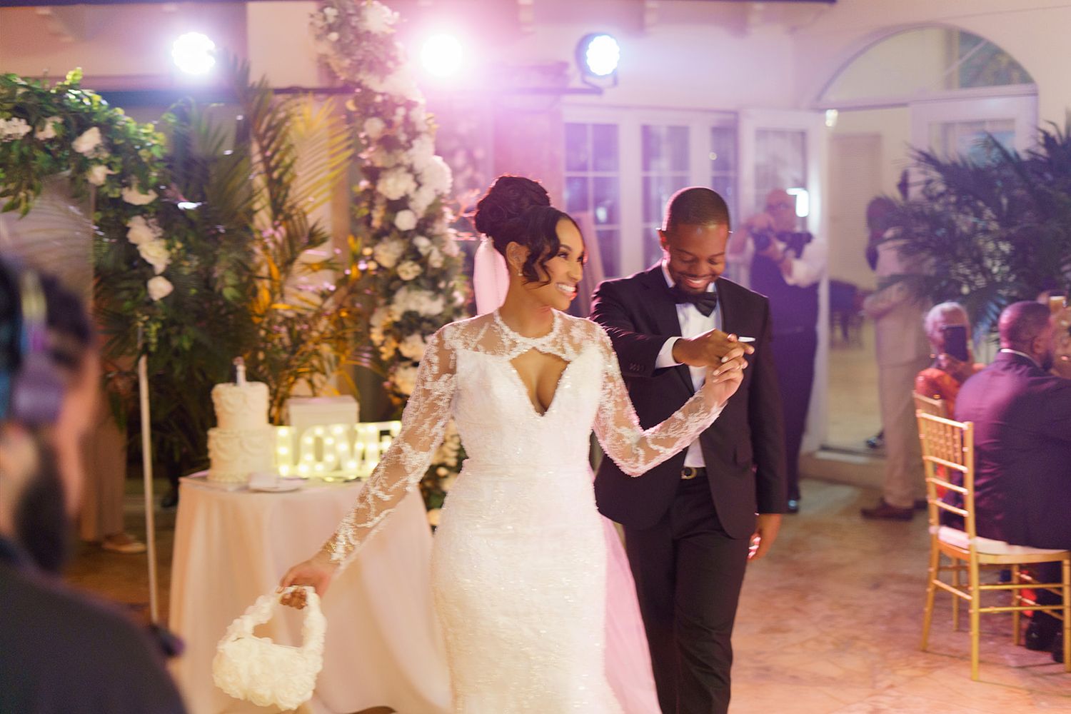 A bride in a lace gown and groom in a tuxedo walk together at an elegant wedding reception.