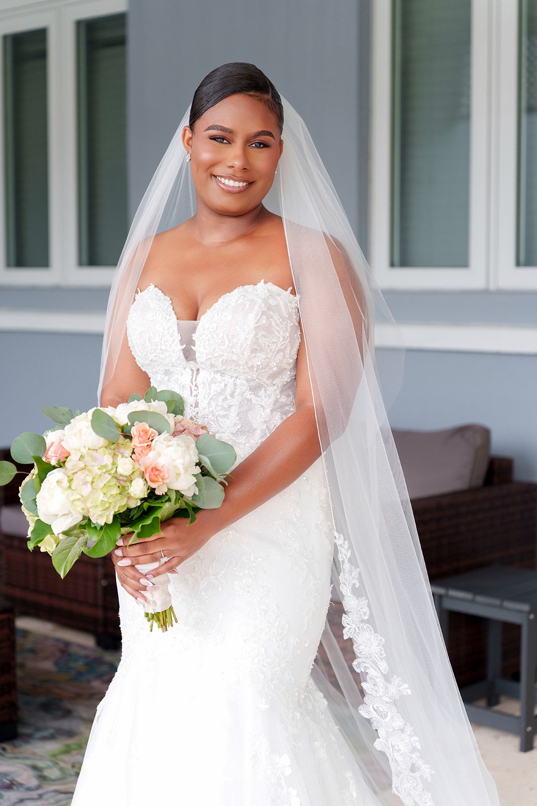 Smiling bride in strapless lace mermaid gown and veil holds peach and white bouquet outdoors.