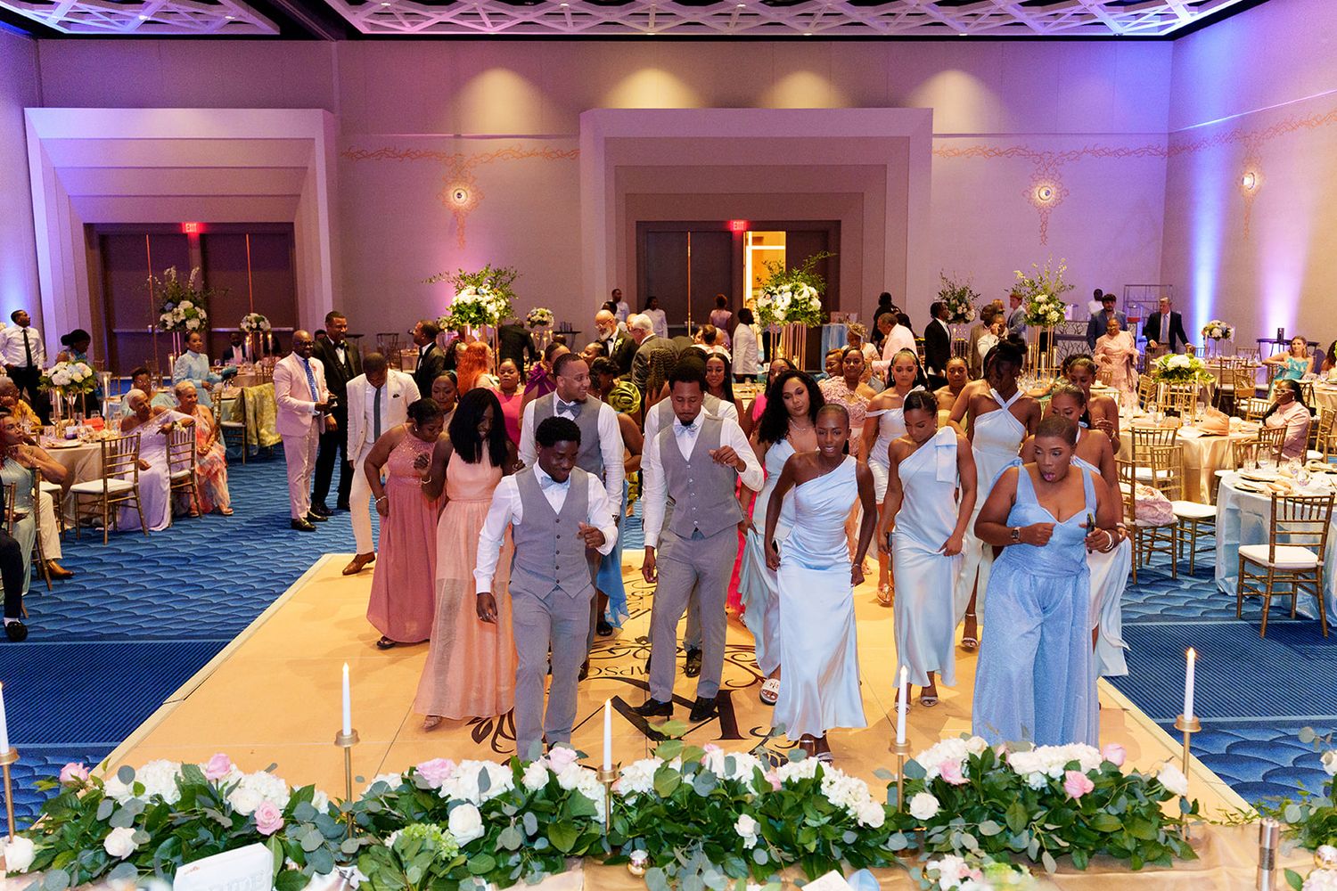 Wedding party processional in grand ballroom with guests seated at decorated tables and floral centerpieces.