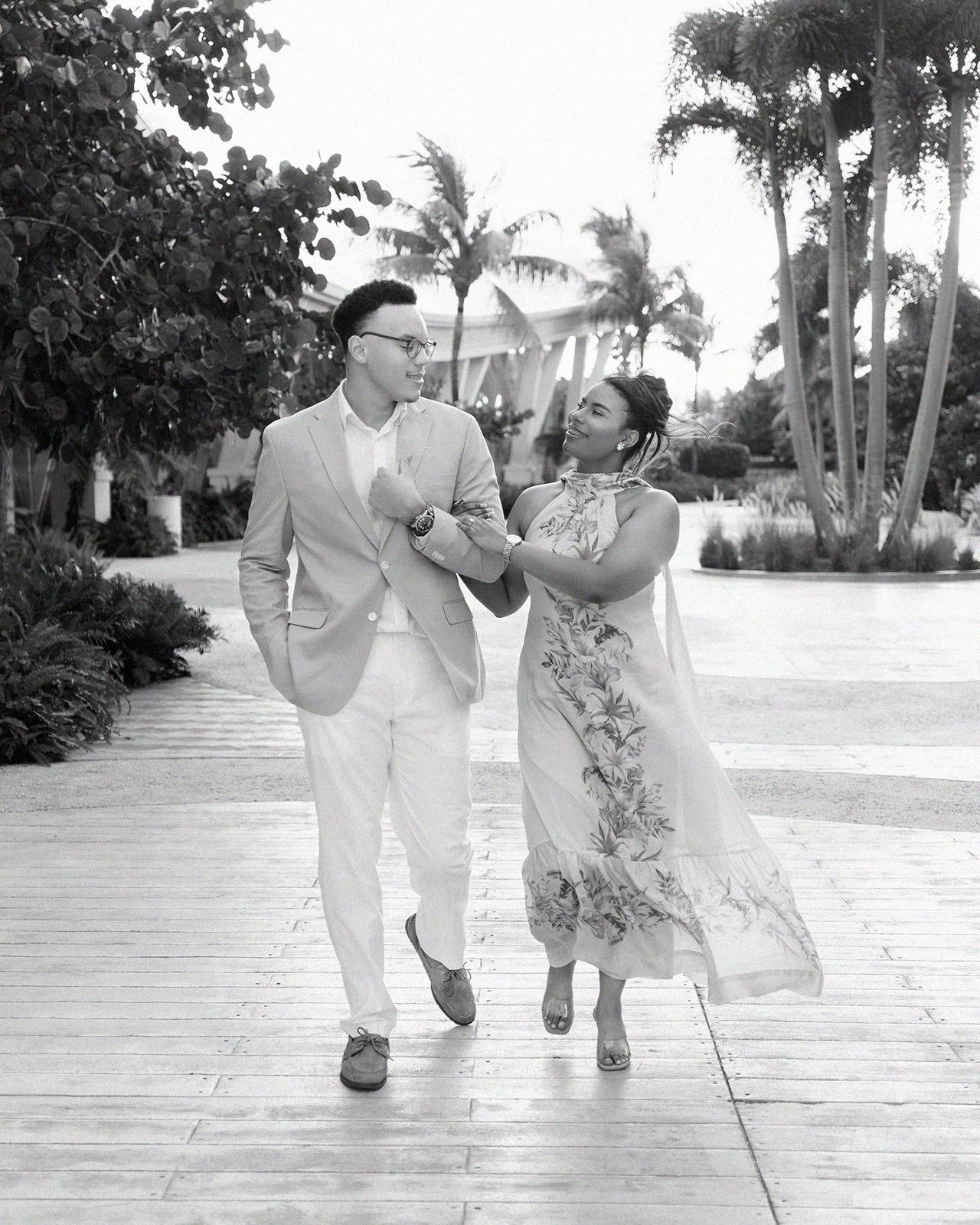 Couple walks hand-in-hand on tropical resort path, smiling at each other, dressed in elegant attire, black and white photo.