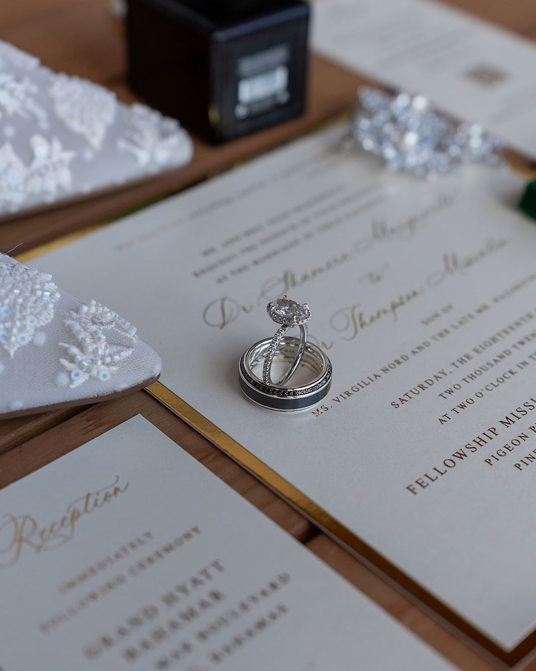 Elegant wedding invitation suite with gold script, diamond engagement ring, and wedding bands displayed on a wooden surface.