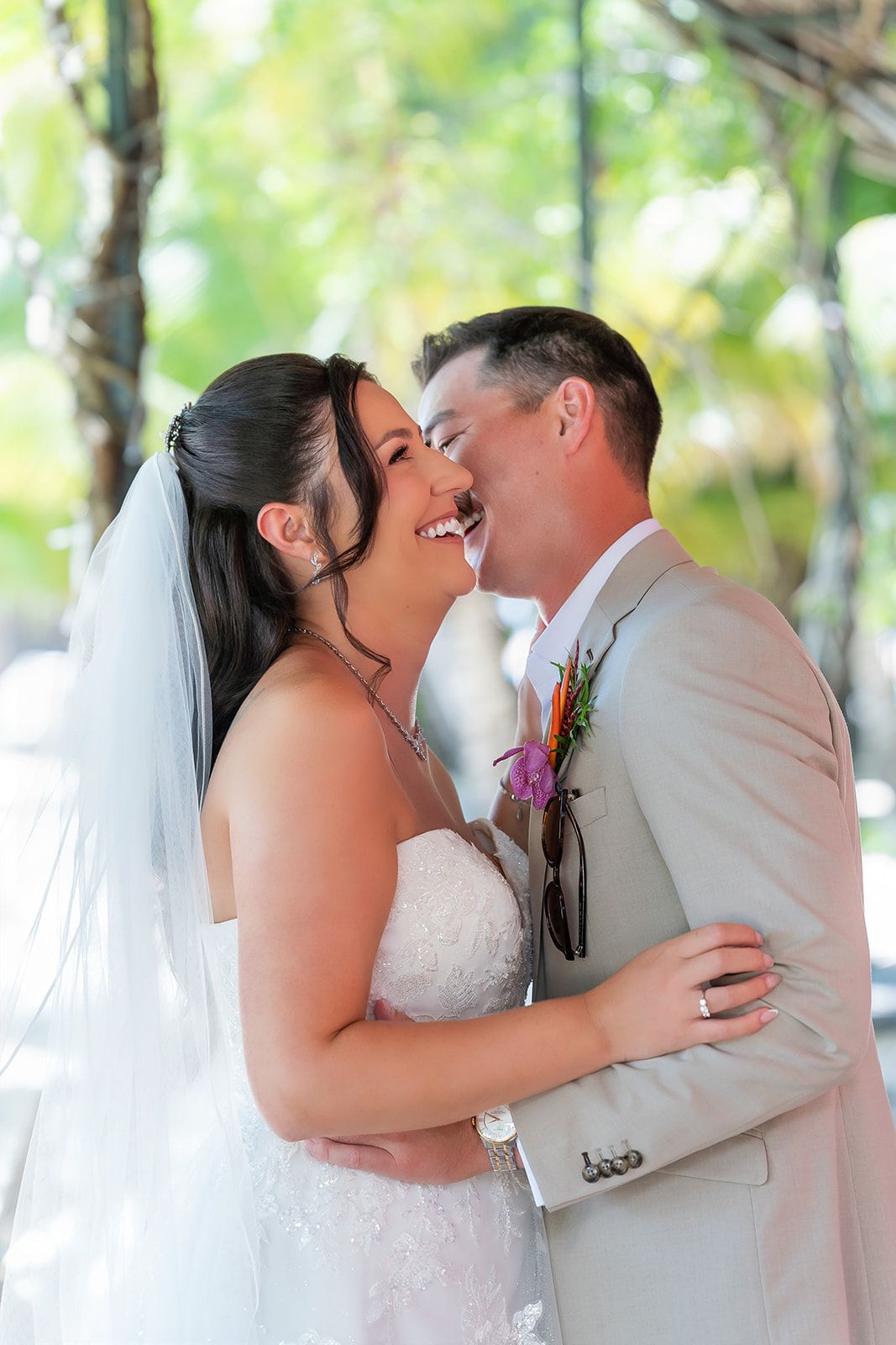 A joyful bride and groom share a laughing embrace outdoors, with the bride in a white gown and veil and groom in a tan suit.