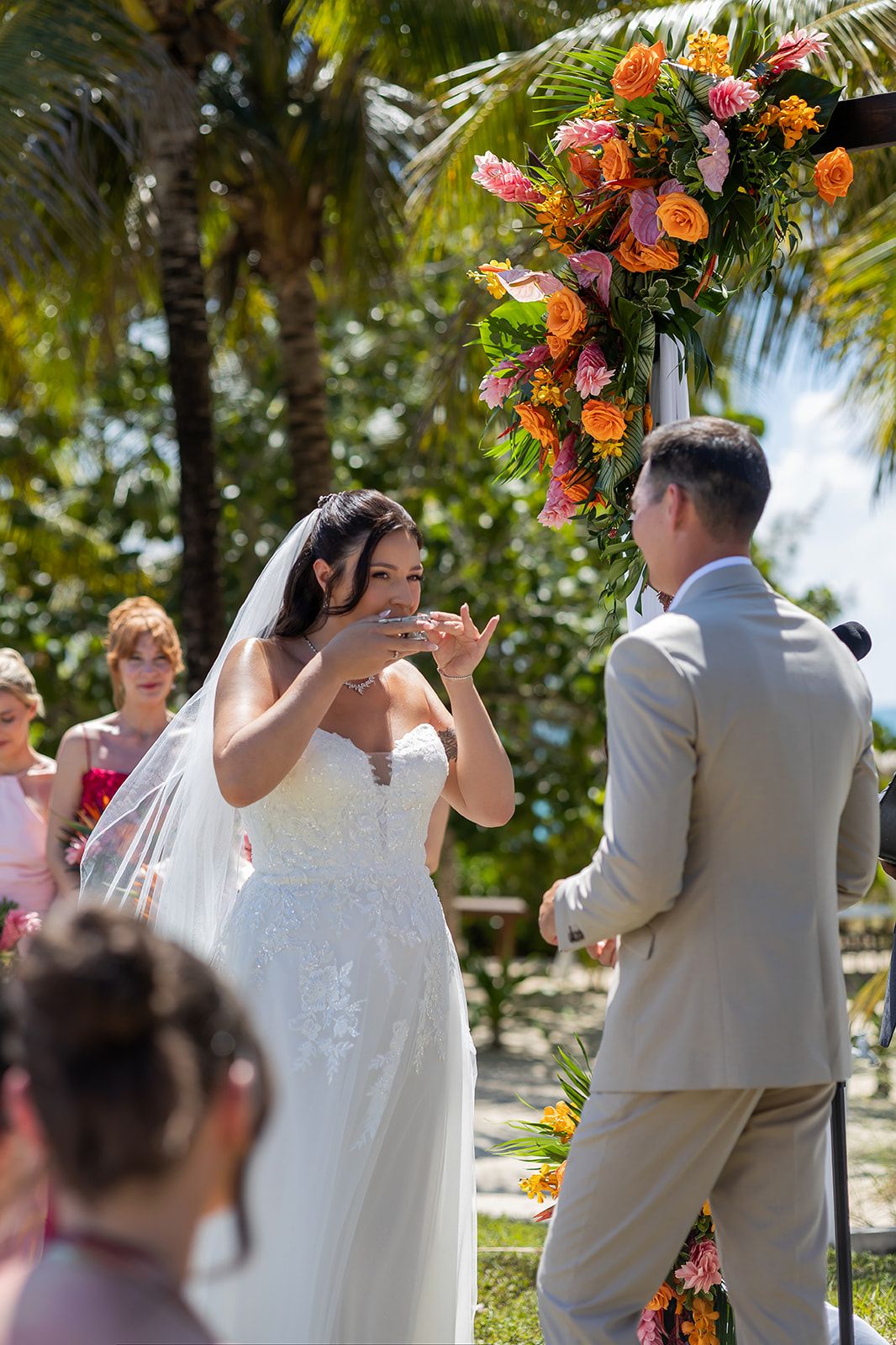 Bride reads vows to groom at tropical outdoor wedding ceremony with vibrant orange and pink floral arch.