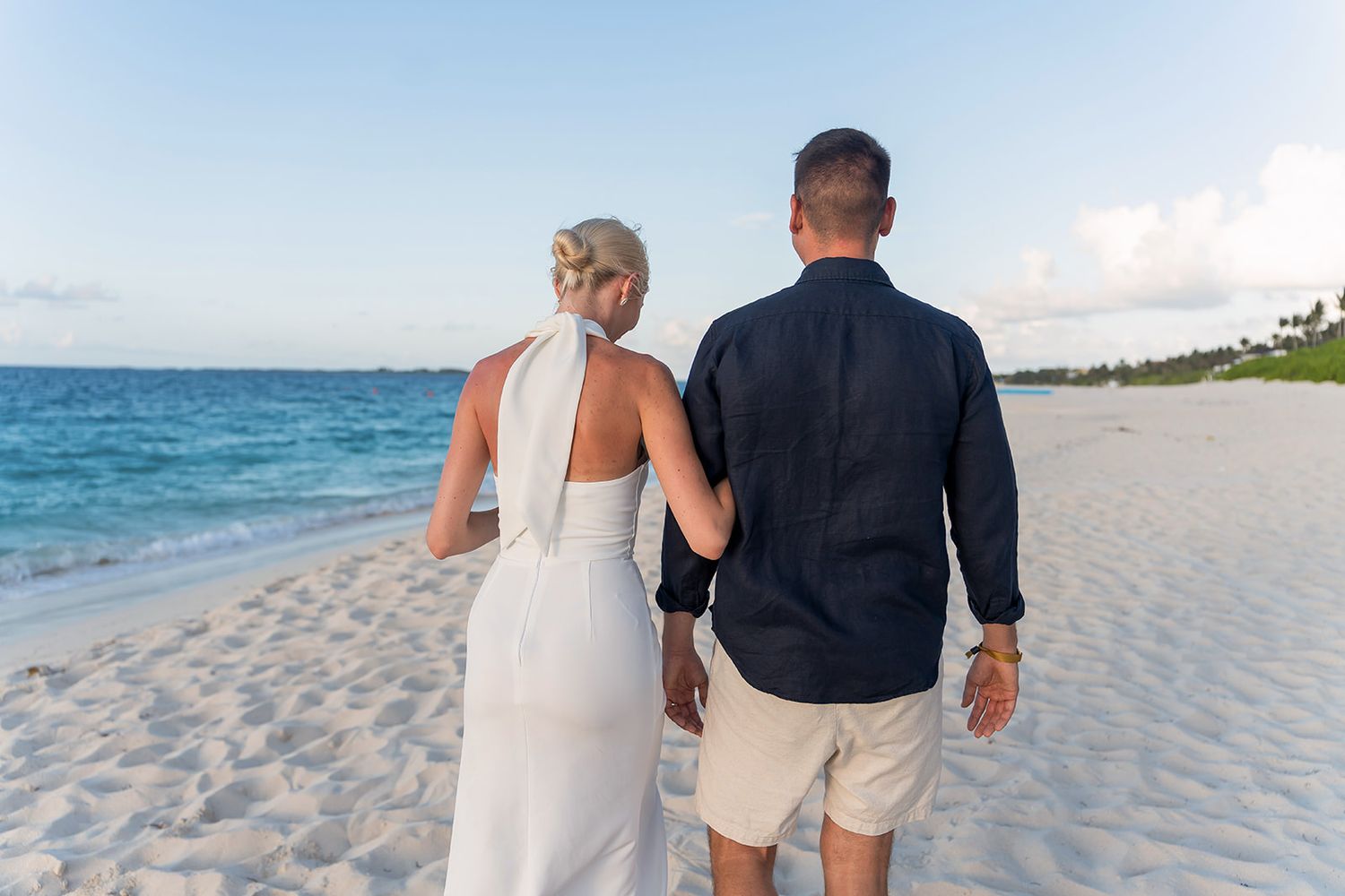 Newlywed couple walking hand in hand on a tropical white sand beach, bride in white gown, groom in navy shirt.