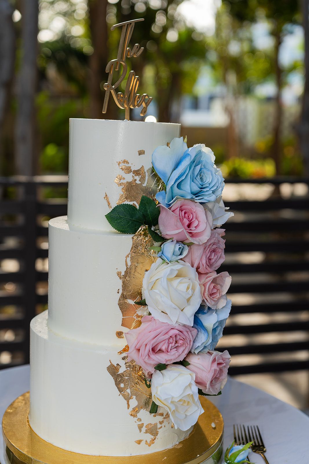 Three-tier white wedding cake with gold accents and colorful roses topped with custom cake topper.