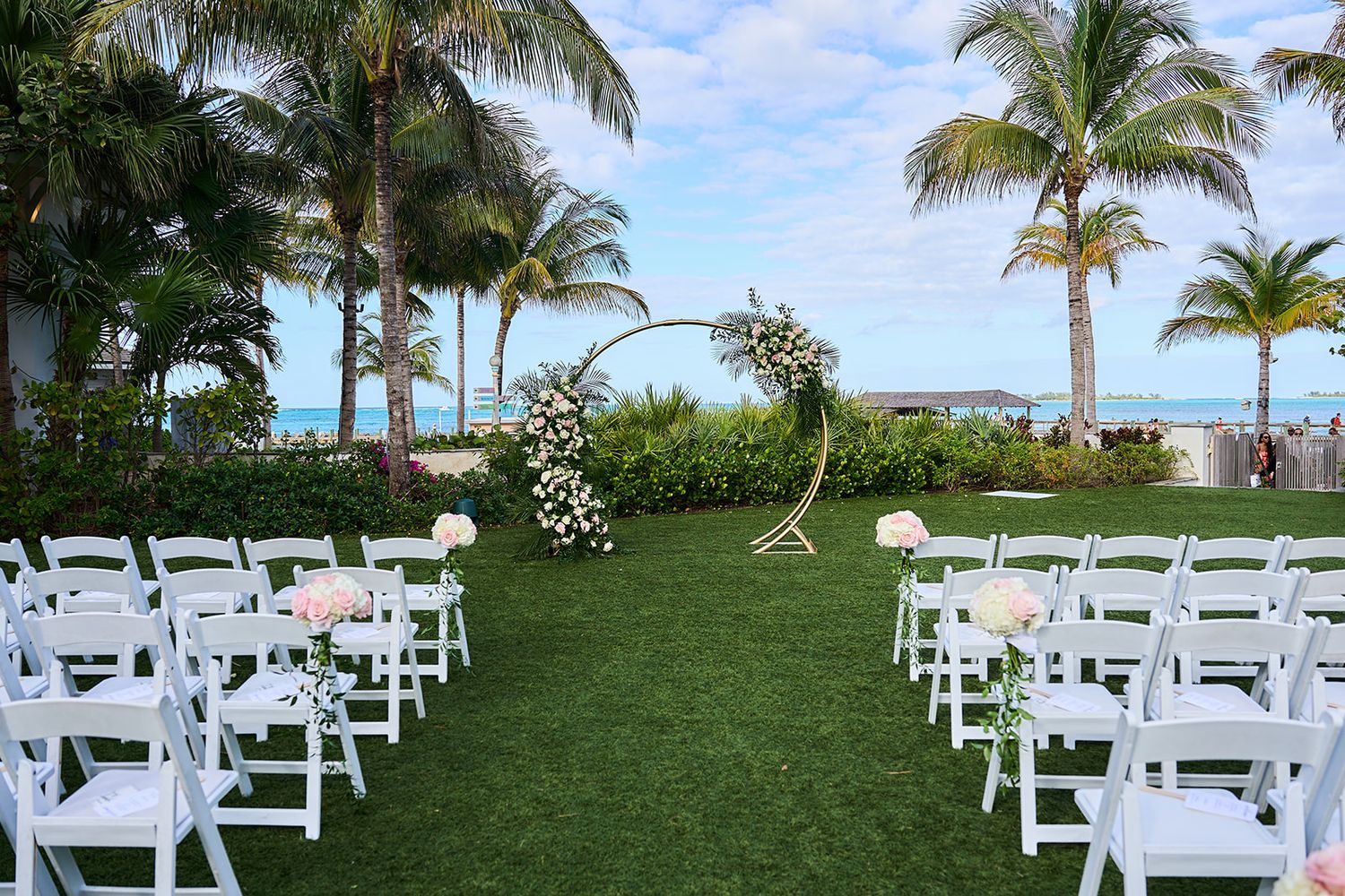 Outdoor beachside wedding ceremony setup with white chairs, floral arch, and palm trees overlooking turquoise ocean waters.