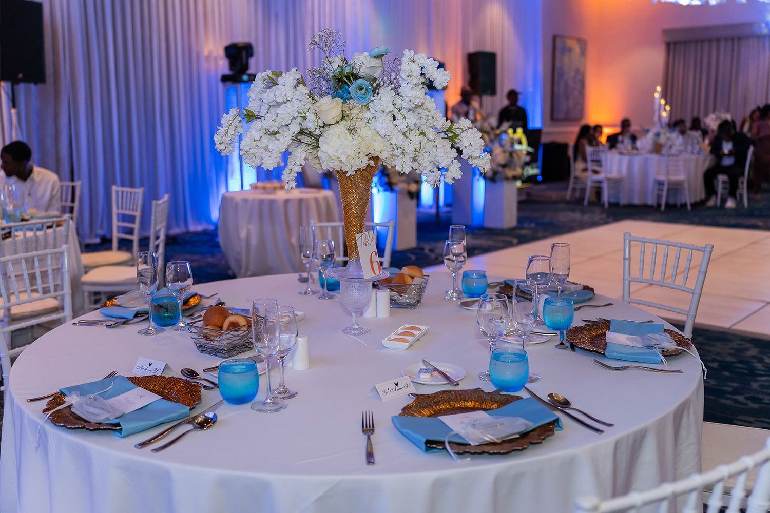 Elegant wedding reception table with white floral centerpiece, blue accents, gold chargers, and Chiavari chairs in a ballroom.