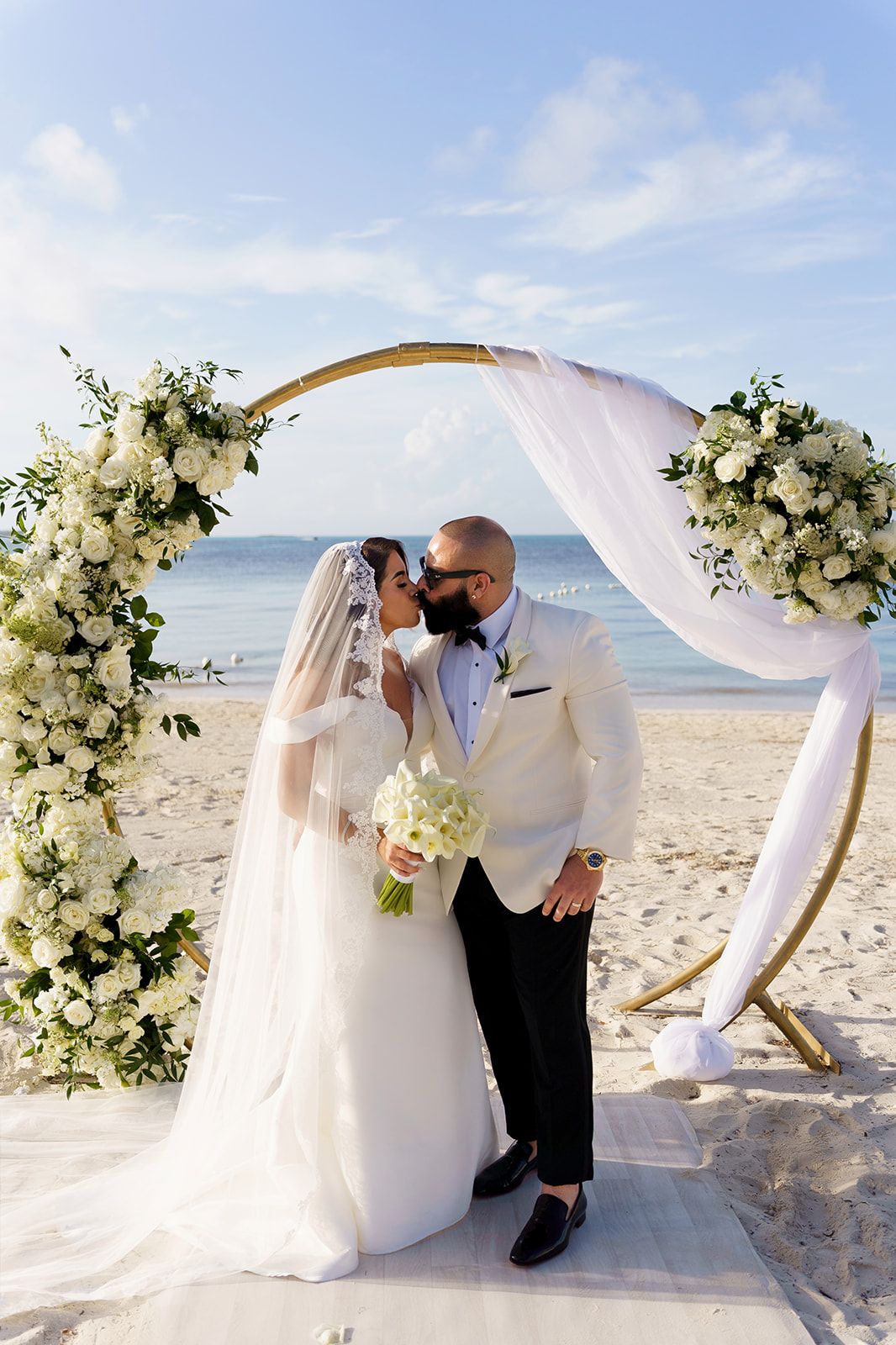 Bride and groom share first kiss under floral arch on beach wedding ceremony with white roses and greenery.