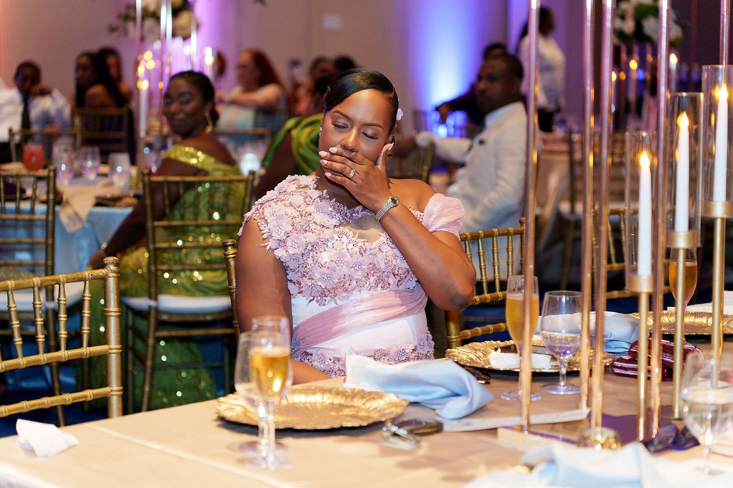 A woman in a lavender lace gown sits at an elegant gold-accented banquet table, looking emotional at a formal event.