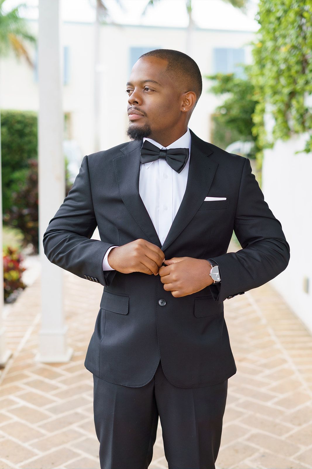 Dapper groom in black tuxedo with bow tie buttoning jacket outdoors at a wedding venue.