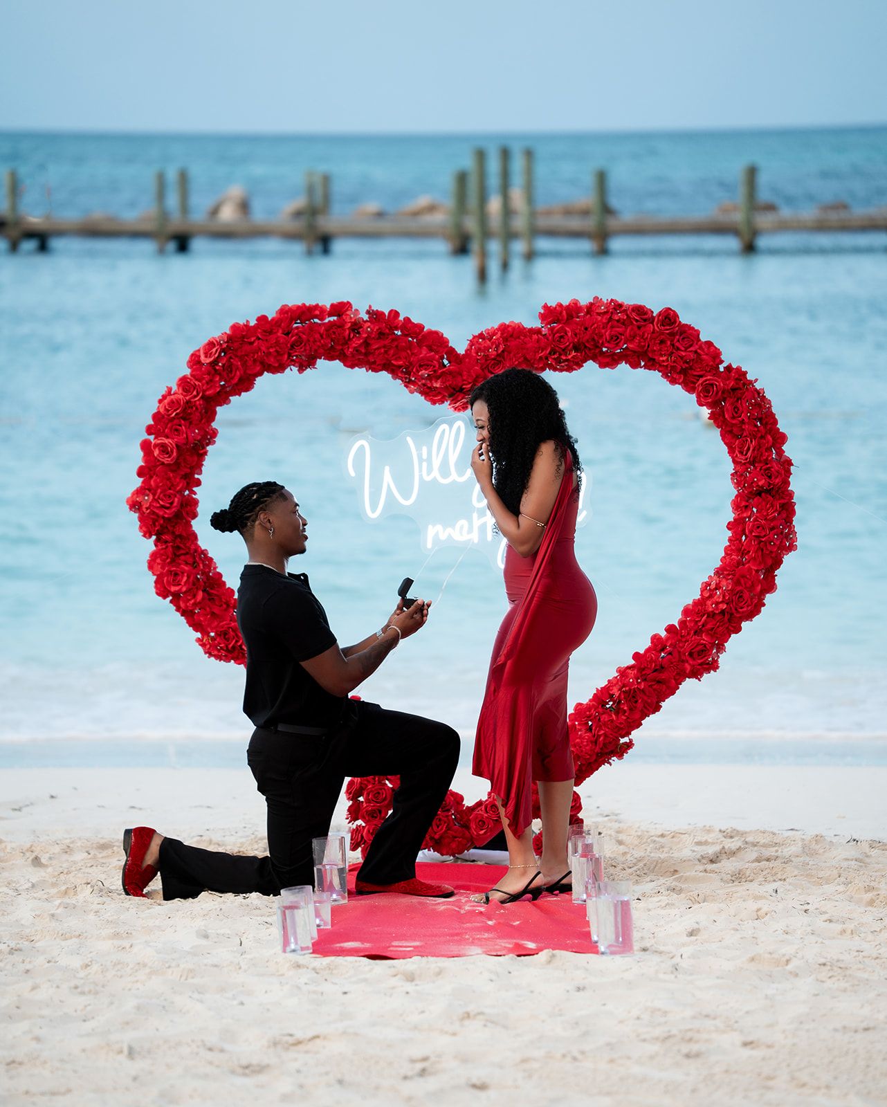 Man proposes to woman on beach with large red rose heart arch and neon sign by turquoise ocean waters.