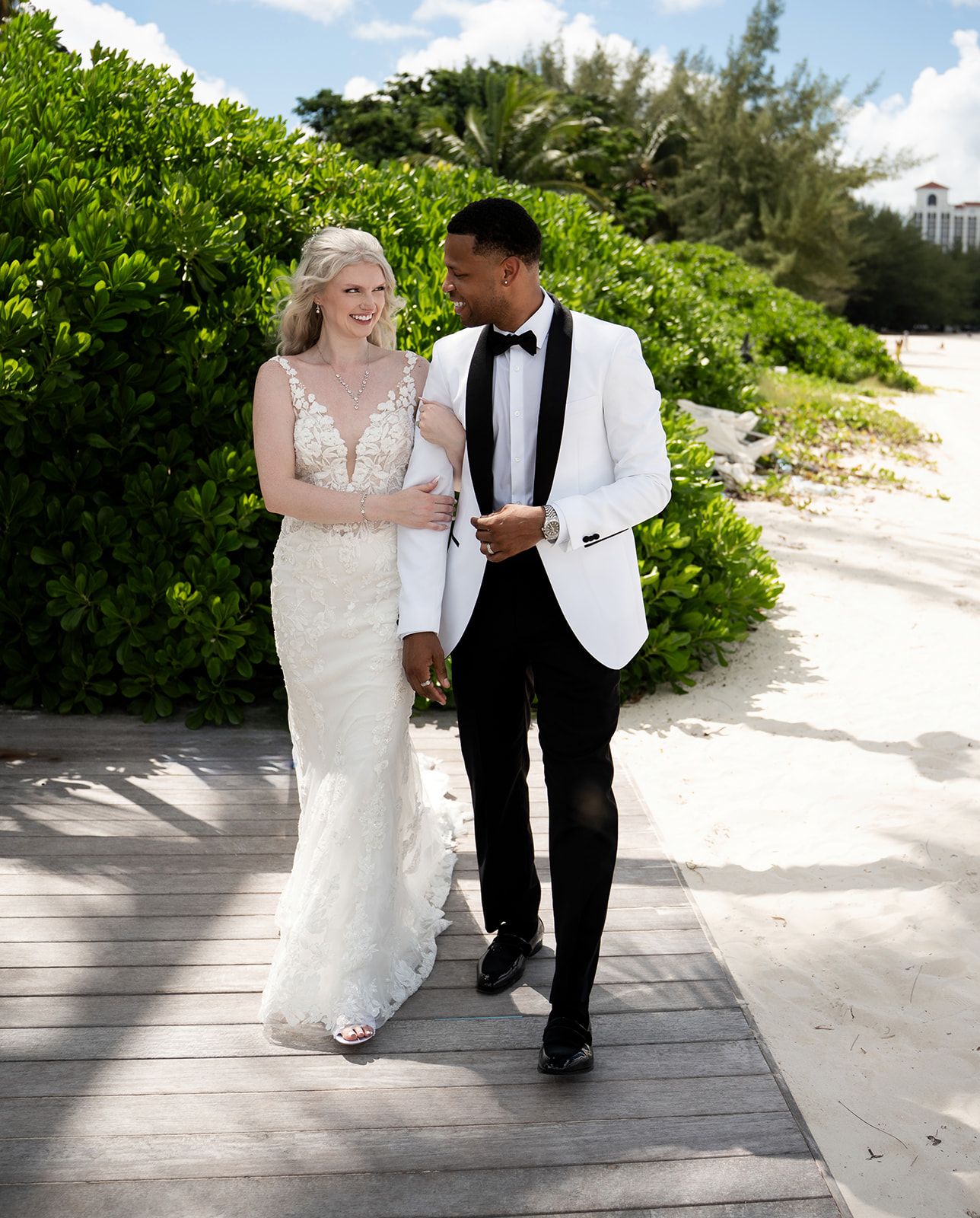 A couple in wedding attire walks on a beachside boardwalk, bride in white gown and groom in white tuxedo jacket.