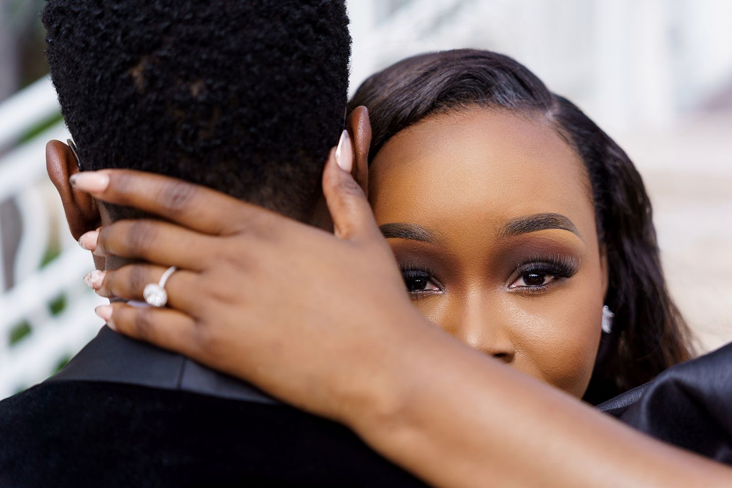 A woman hugs a man while showing off her diamond engagement ring, with elegant makeup and pearl earrings.