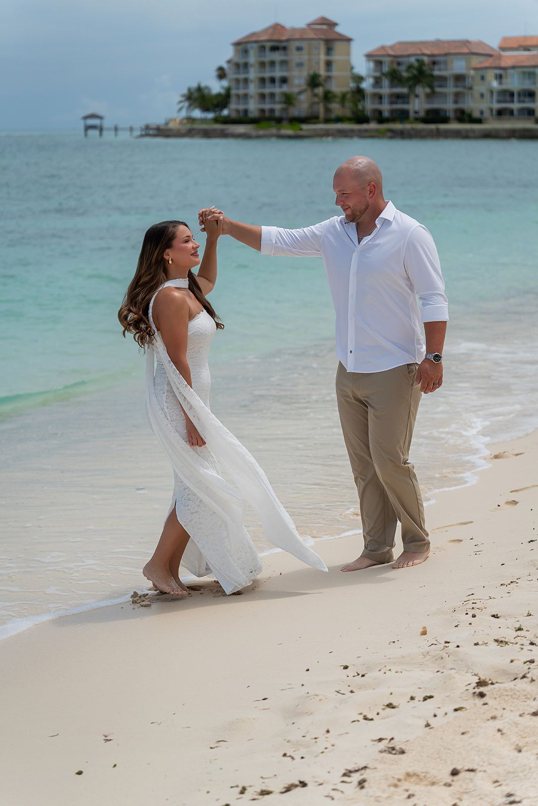 Groom twirling bride in white dress on tropical beach with turquoise water.