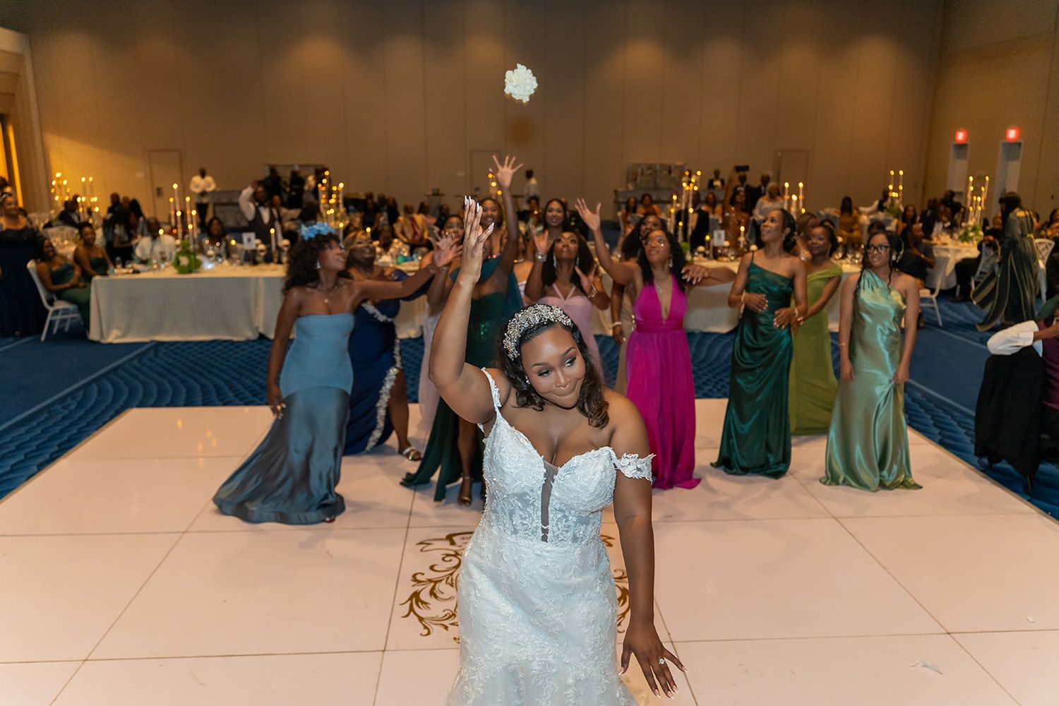 Bride in white lace gown tosses bouquet on dance floor surrounded by bridesmaids in colorful gowns at elegant wedding reception.