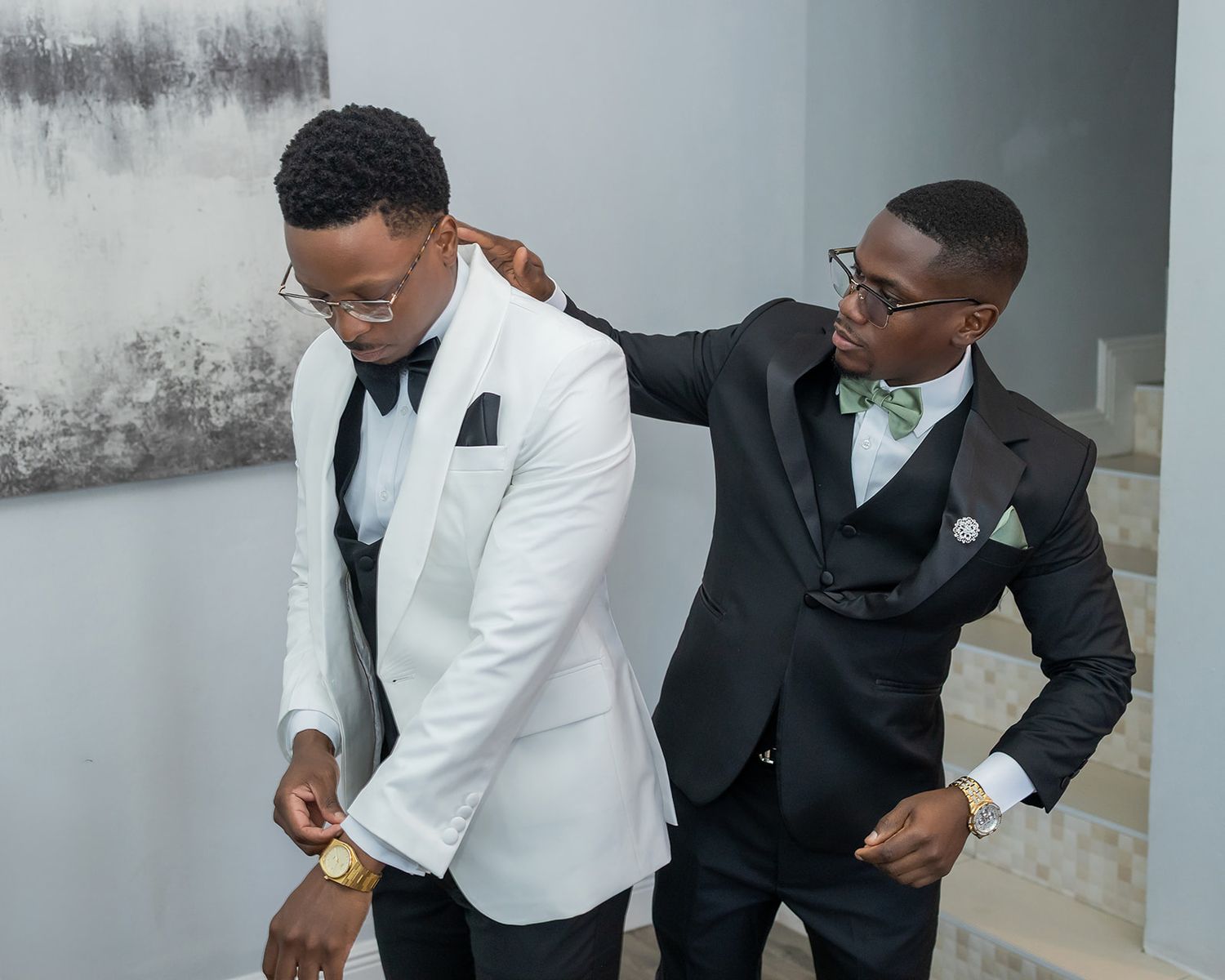 Two men in formal suits, one in white tuxedo and one in black suit with green bow tie, at an elegant event.