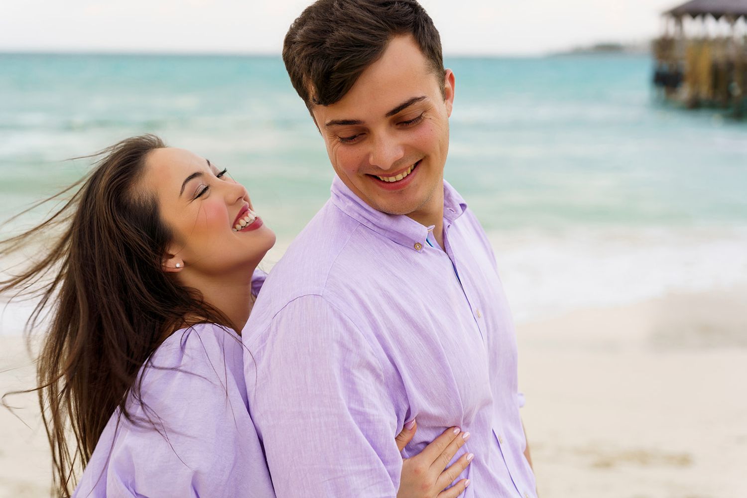 Happy couple in matching lavender shirts embracing and laughing on a sunny beach with turquoise ocean waves.