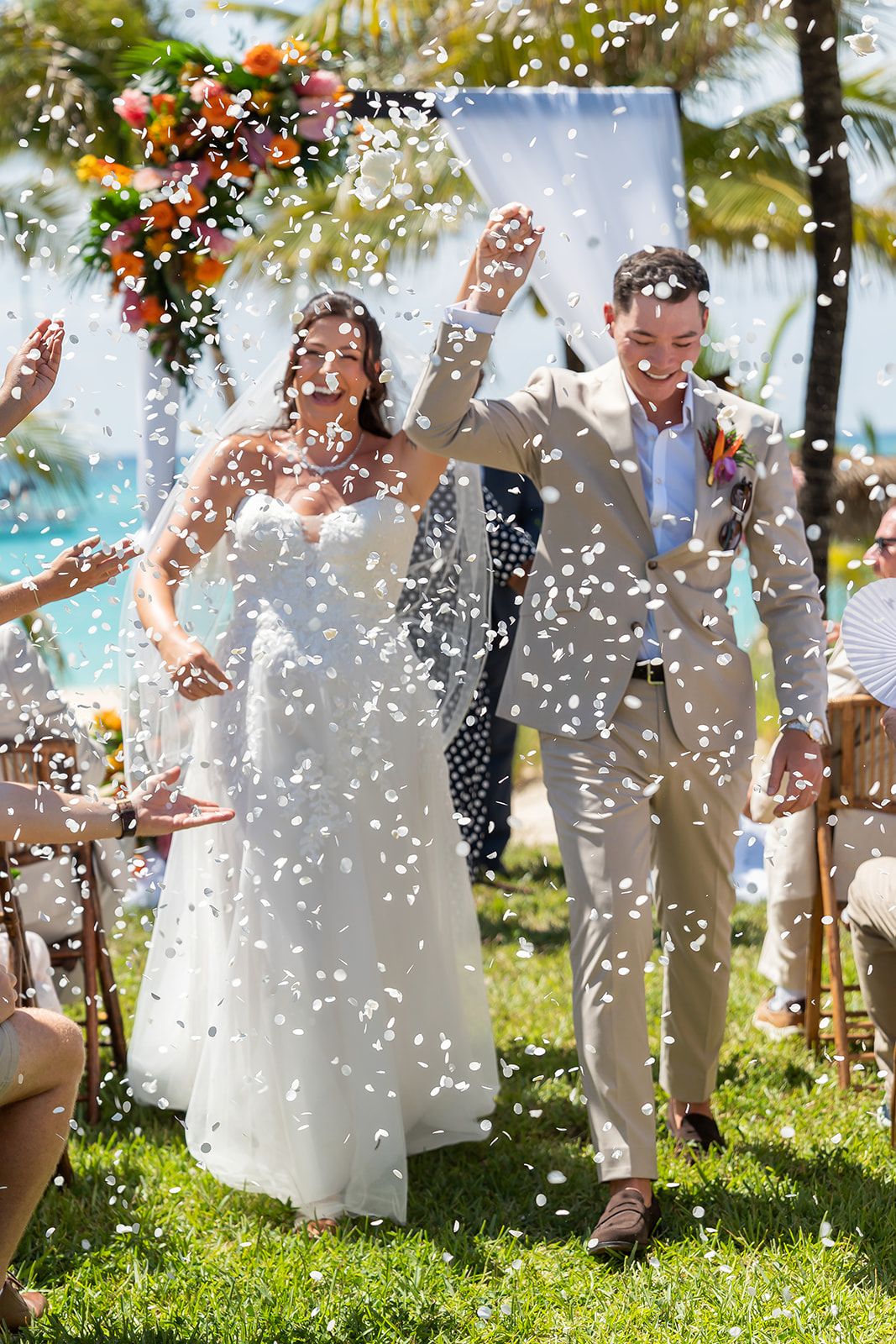 Joyful newlyweds walk down the aisle as white confetti fills the air at an outdoor beach wedding ceremony.