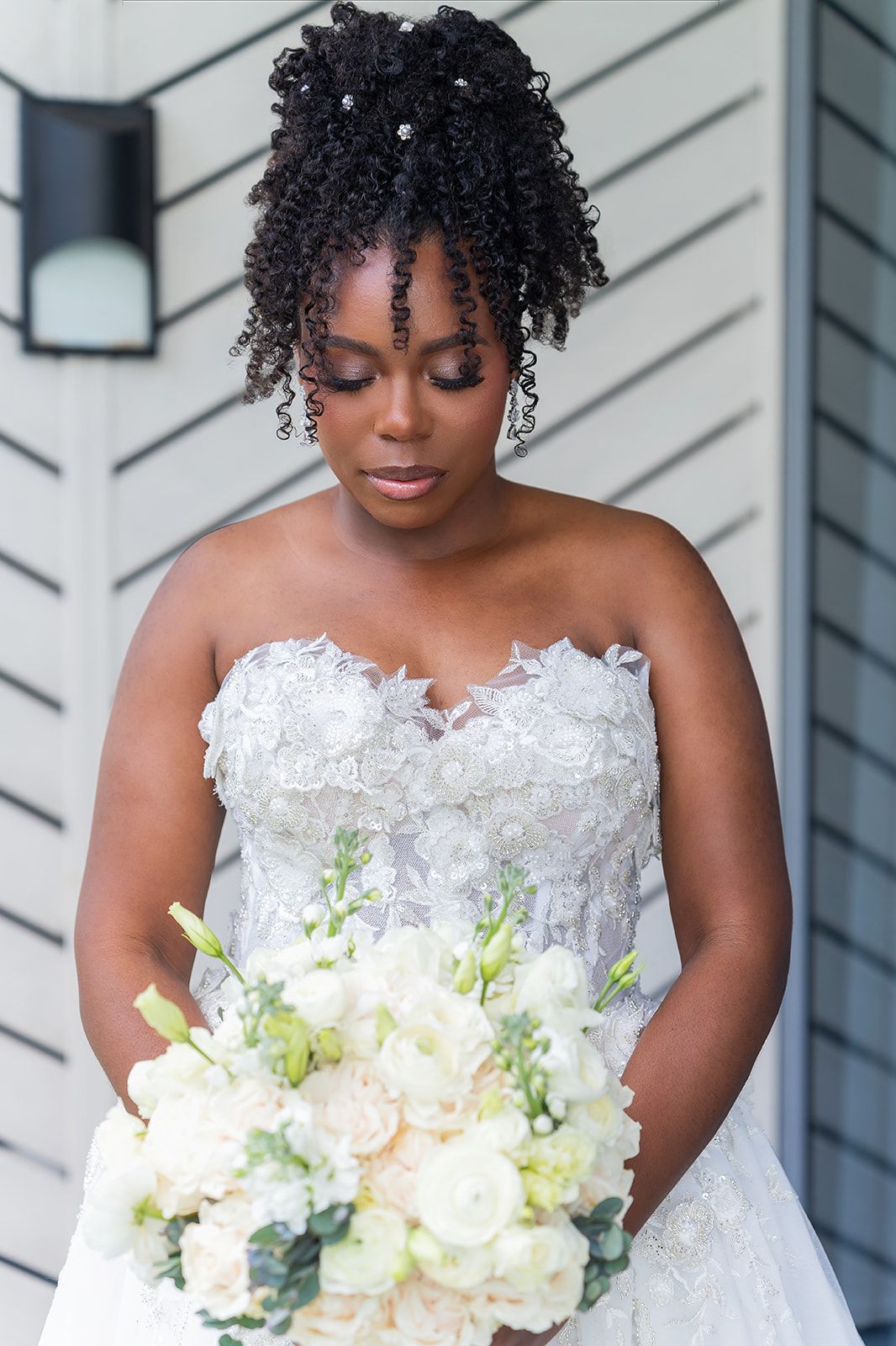 Beautiful Black bride in strapless lace ball gown holding white and blush floral bouquet with eyes downcast.