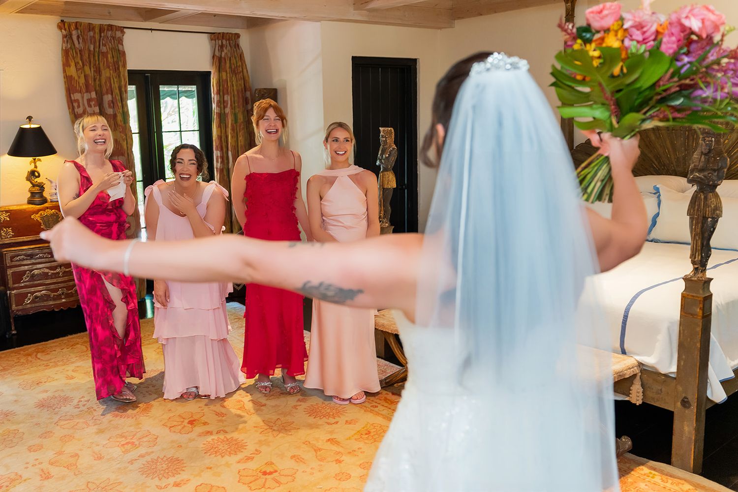 Bride in white gown and veil shows bouquet to bridesmaids in pink and red dresses in elegant hotel room.
