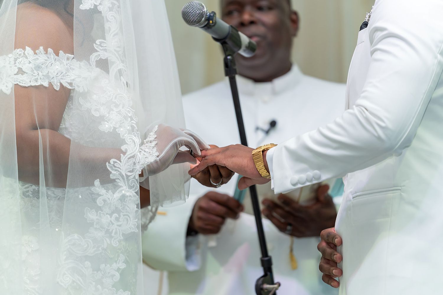 A groom places a wedding ring on the bride's finger during a ceremony officiated by a minister in white.