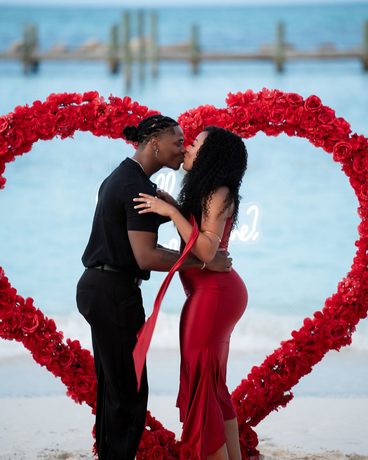Couple kissing in front of a large red rose heart arch on a beach, woman in red dress.