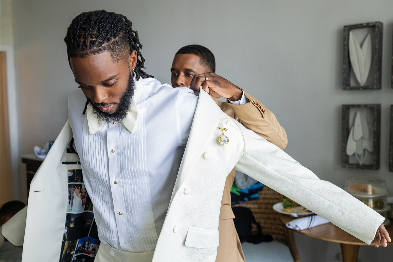 A groomsman helps a groom put on a white suit jacket while preparing for a wedding ceremony.