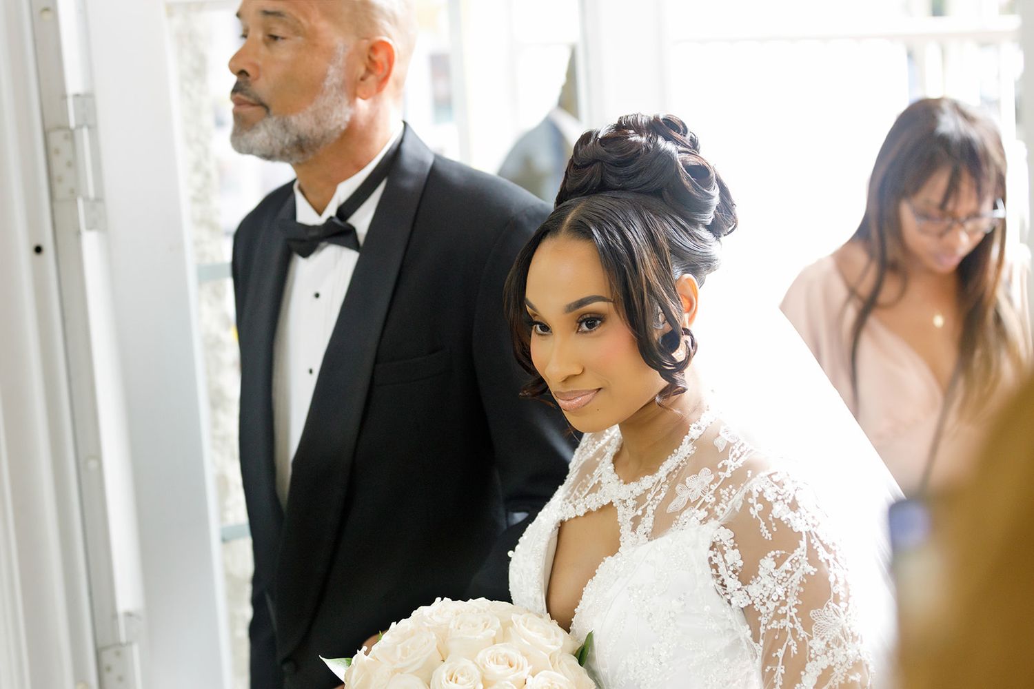Bride in lace wedding gown holding white bouquet smiles beside man in tuxedo at elegant wedding ceremony.