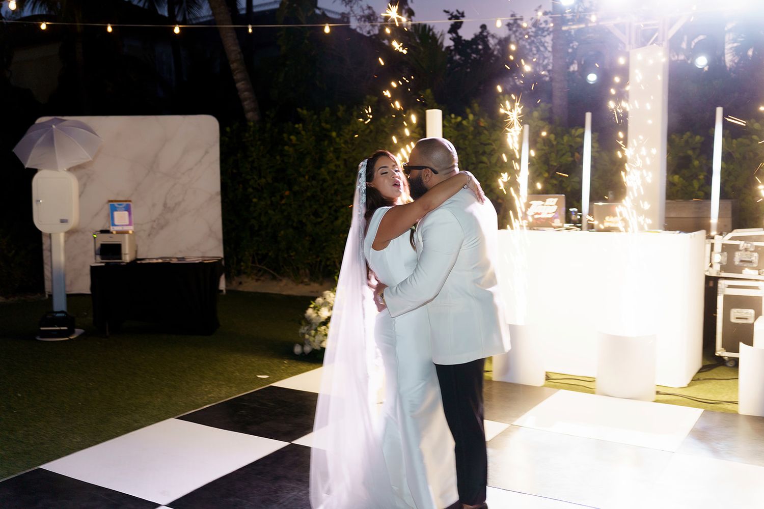 Bride and groom share first dance on checkered floor with sparklers at outdoor wedding reception.