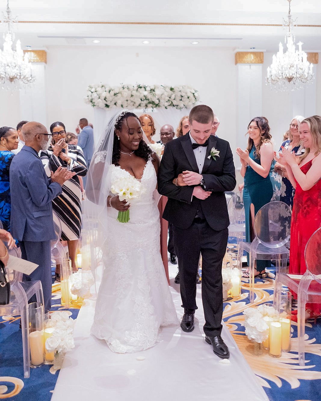 Newlywed couple walks down the aisle after wedding ceremony in elegant ballroom with candles and cheering guests.