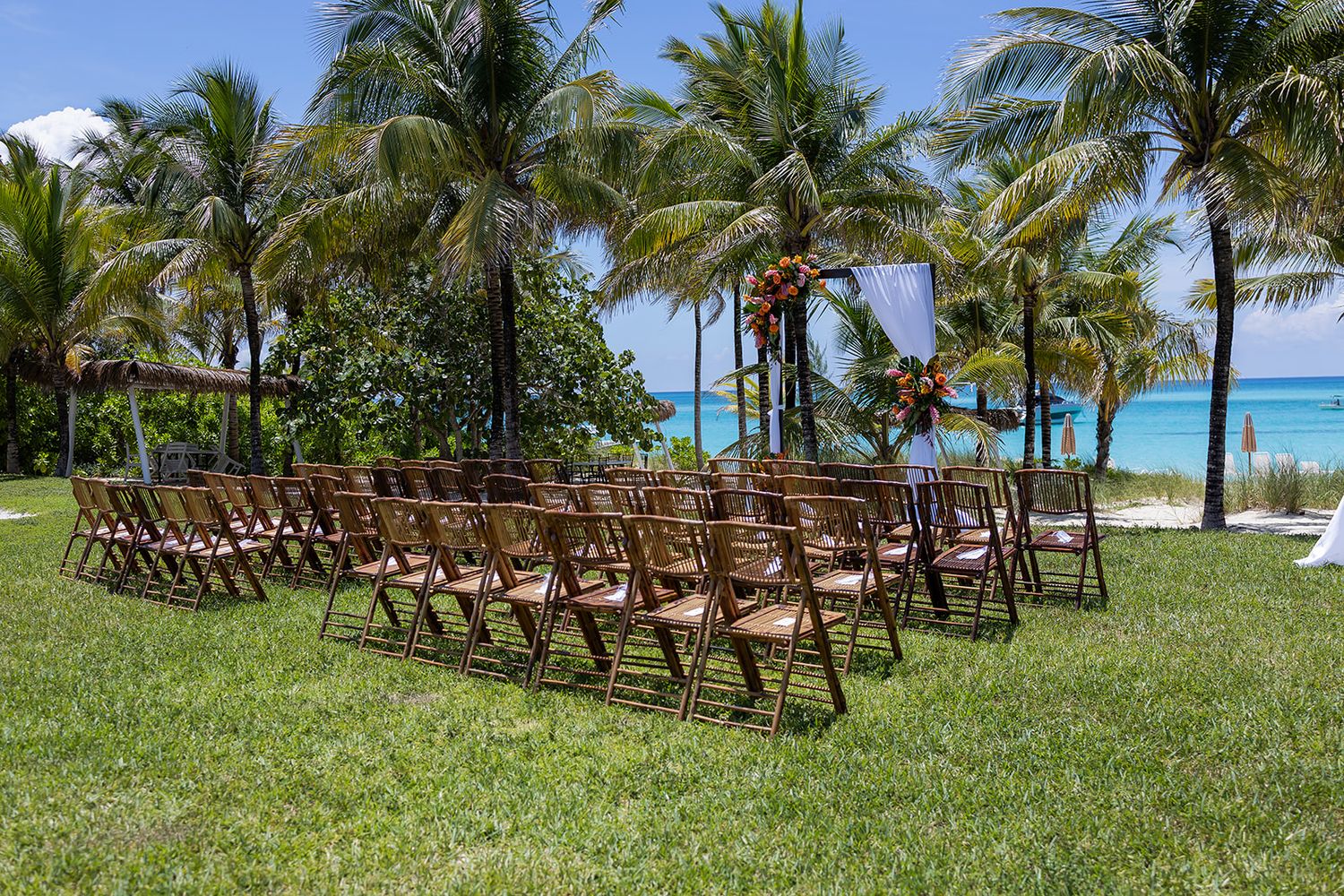 Outdoor beach wedding ceremony setup with wooden chairs on green lawn surrounded by palm trees and ocean view.