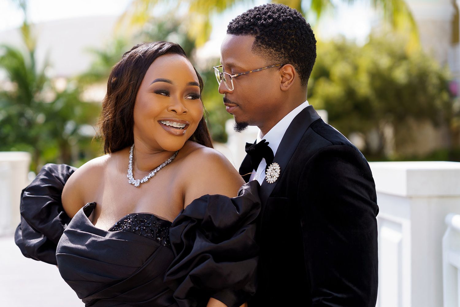 Elegant couple in formal black attire posing outdoors, woman in off-shoulder gown and man in tuxedo with boutonniere.